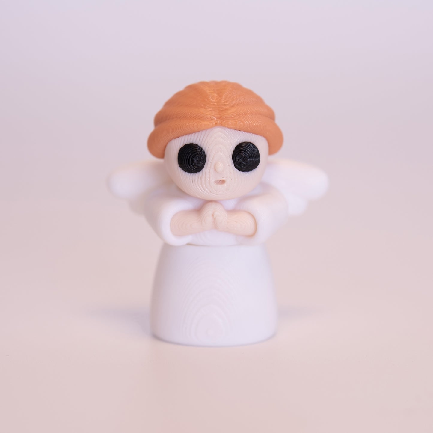 A 3D printed standing Angel figurine with her hands together praying. The Angel has beige skin, round black eyes, orange hair, a white dress and white wings on an off white background.