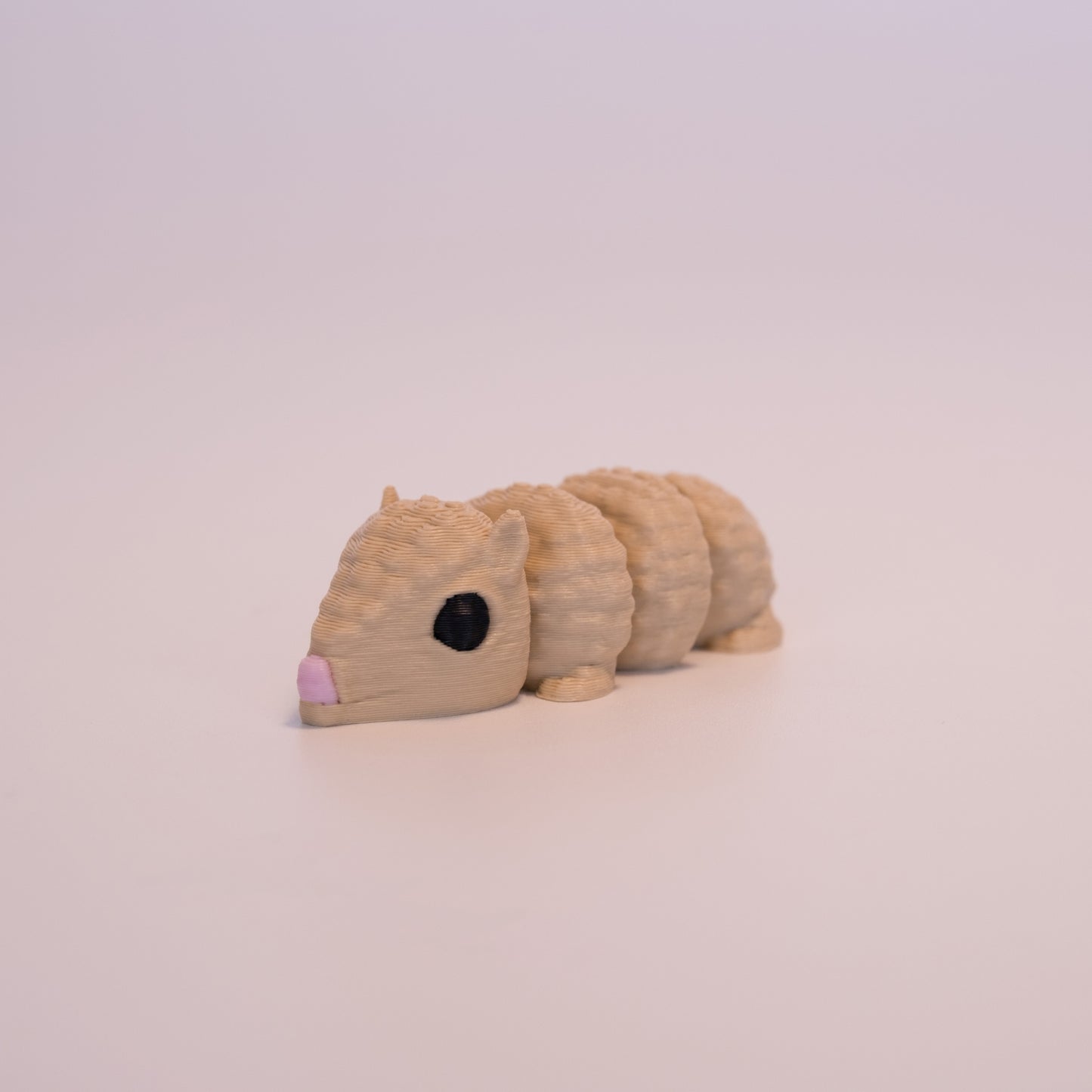 A 3D printed Armadillo figurine on an off white background. The figurine is mostly tan with round black eyes and a pink nose.