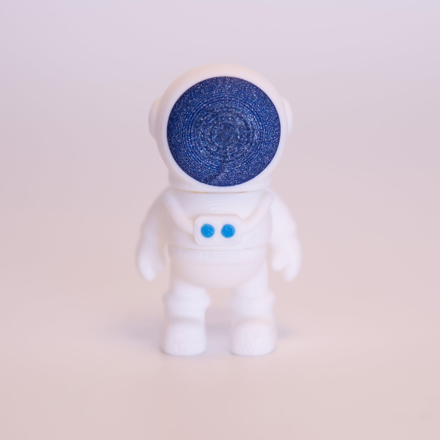 A 3D printed Astronaut figurine standing on an off white background. The Astronaut suit is all white, with the face of the helmet being shiny blue, with silver sparkles, and two buttons on the suit being light blue.