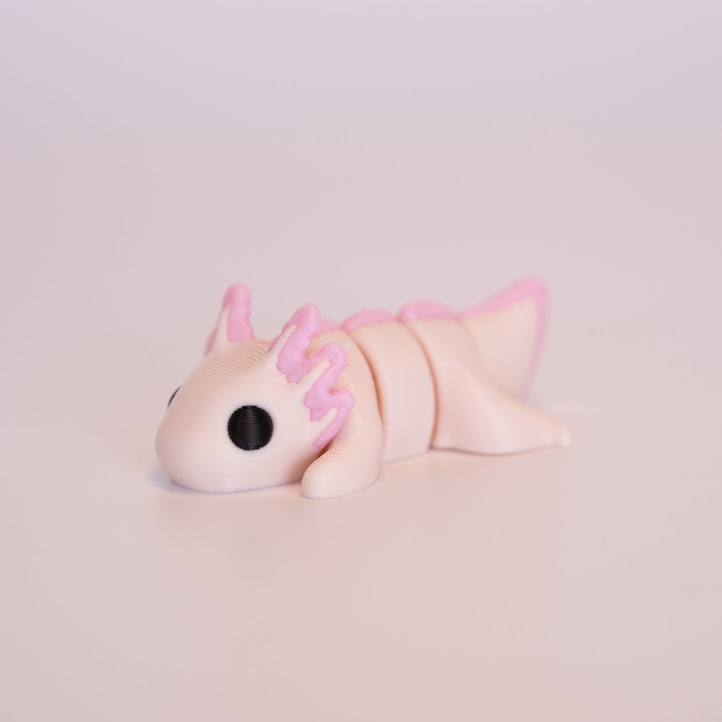 A 3D printed Axolotl figurine laying on its stomach over an off white background. The figurine is mostly light pink with round black eyes and pink accents.