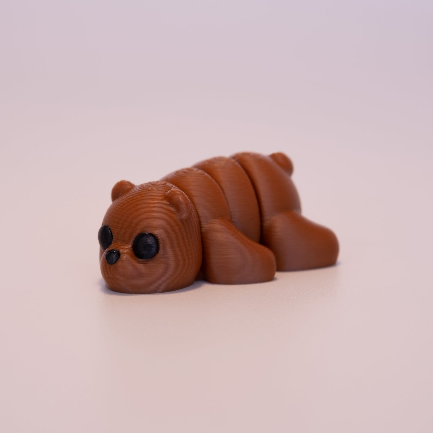 A 3D printed Brown Bear figurine laying on its stomach over an off white background. The figurine is entirely a warm brown with round black eyes and a black button nose.