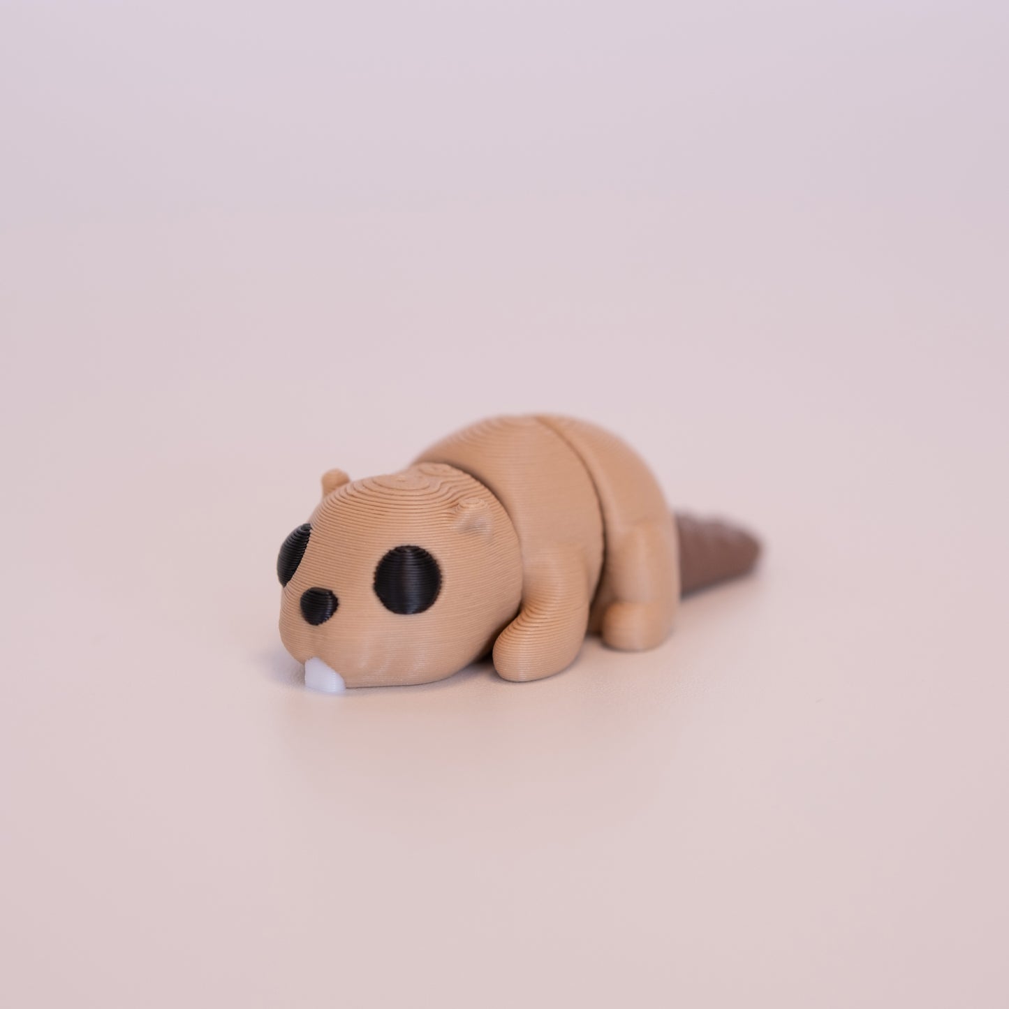 A 3D printed Beaver figurine laying on its stomach over an off white background. The figurine is mostly tan with round black eyes, white buck teeth, and a brown tail.