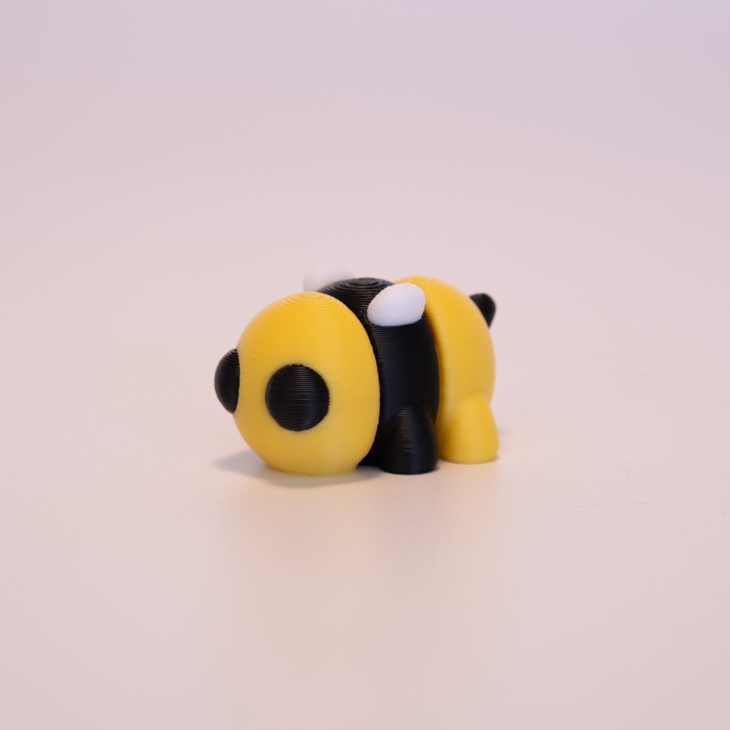 A 3D Printed Bee figurine standing on its 4 legs on an off white background. The Bee’s body has alternating colors of yellow and black, with round black eyes, white wings, and a black stinger.