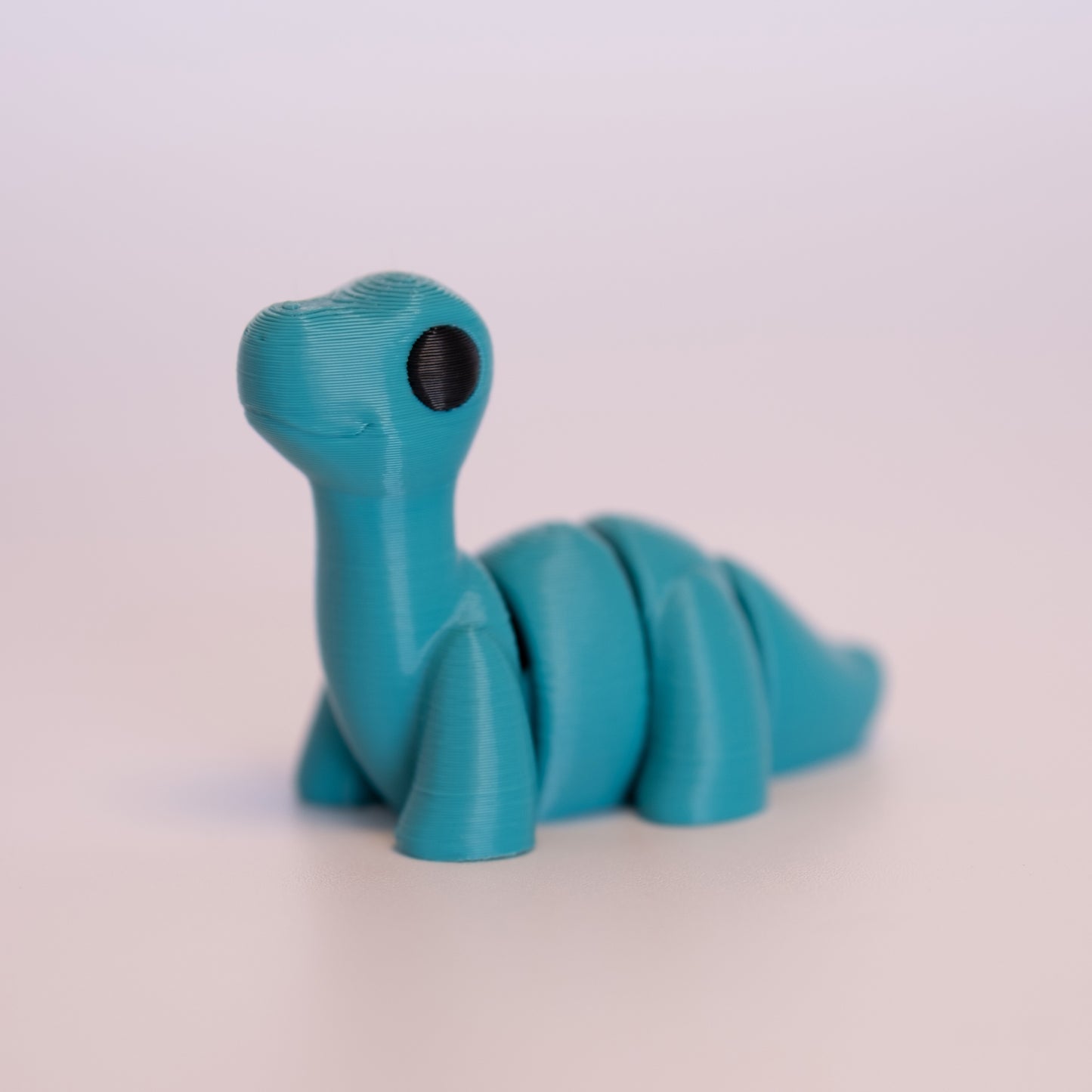 A 3D Printed Brontosaurus figurine standing on its 4 legs on an off white background. The Brontosaurus is entirely teal with round black eyes.