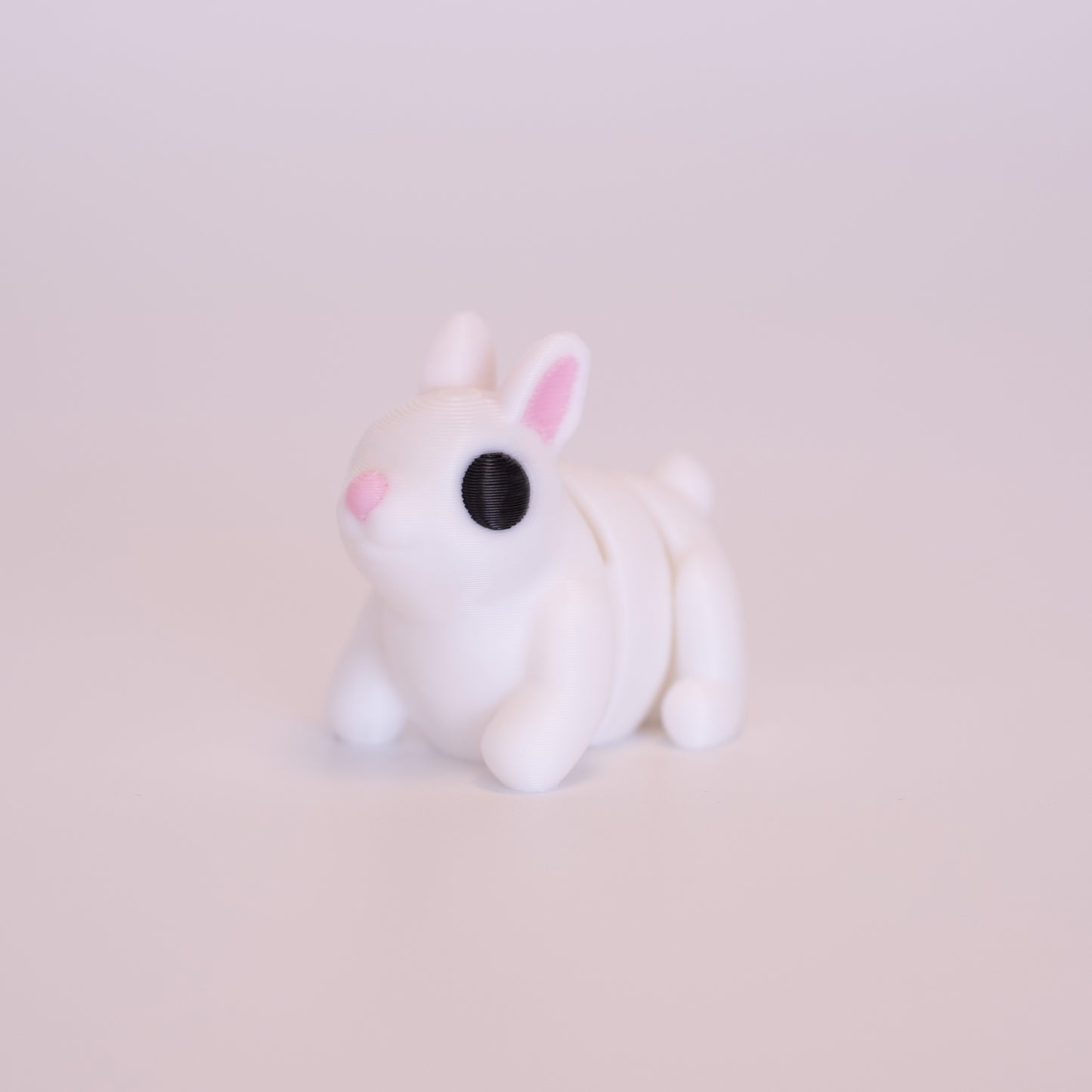 A 3D Printed Bunny figurine standing on its 4 legs on an off white background. The Bunny’s is mostly white with round black eyes, pink nose, and pink accents on the inner parts of its ears.