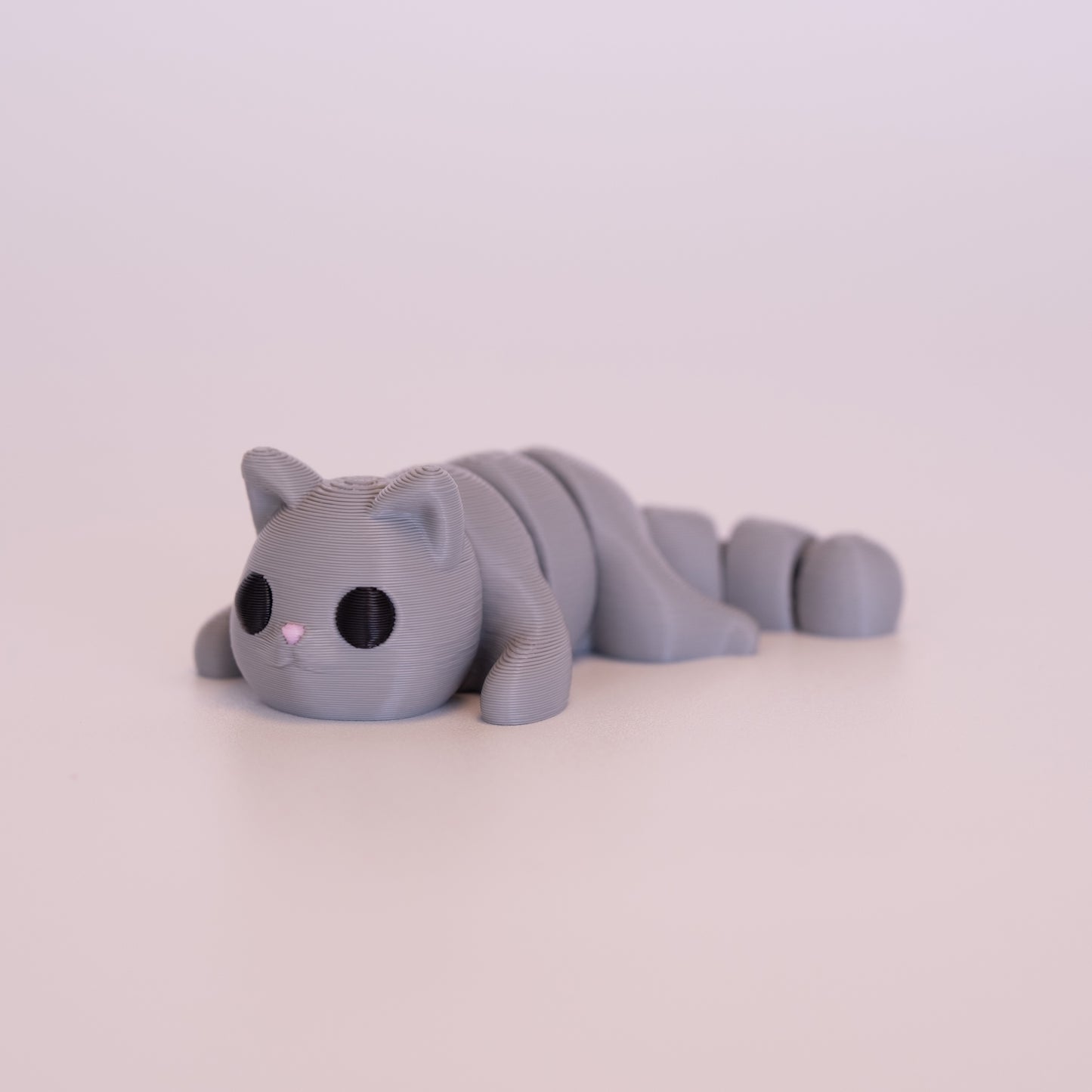 A 3D printed Cat figurine laying on its stomach over an off white background. The figurine is entirely grey with round black eyes and a pink nose.