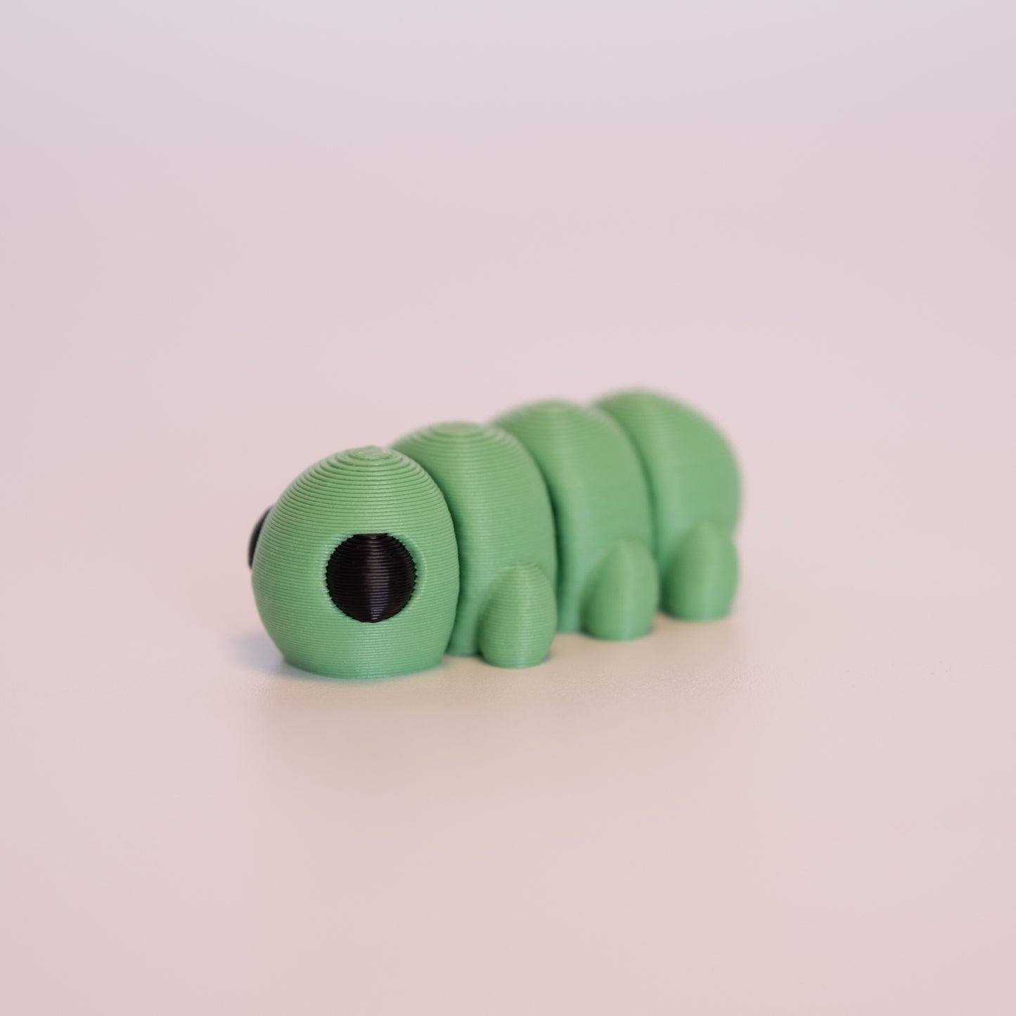 A 3D printed Caterpillar figurine laying flat on an off white background. The figurine is entirely green with round black eyes.