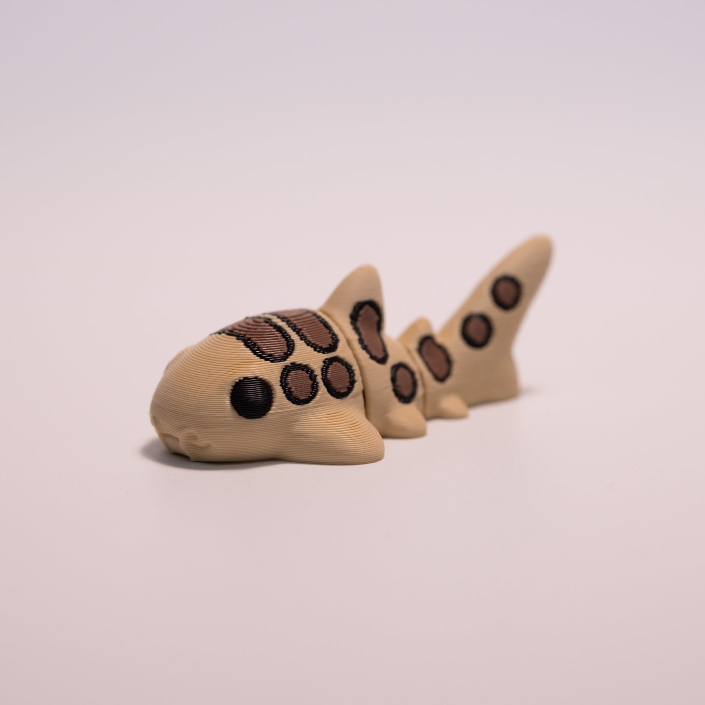 A 3D printed Catshark figurine laying on its stomach over an off white background. The figurine is mostly tan with round black eyes and brown and black patterned accents.