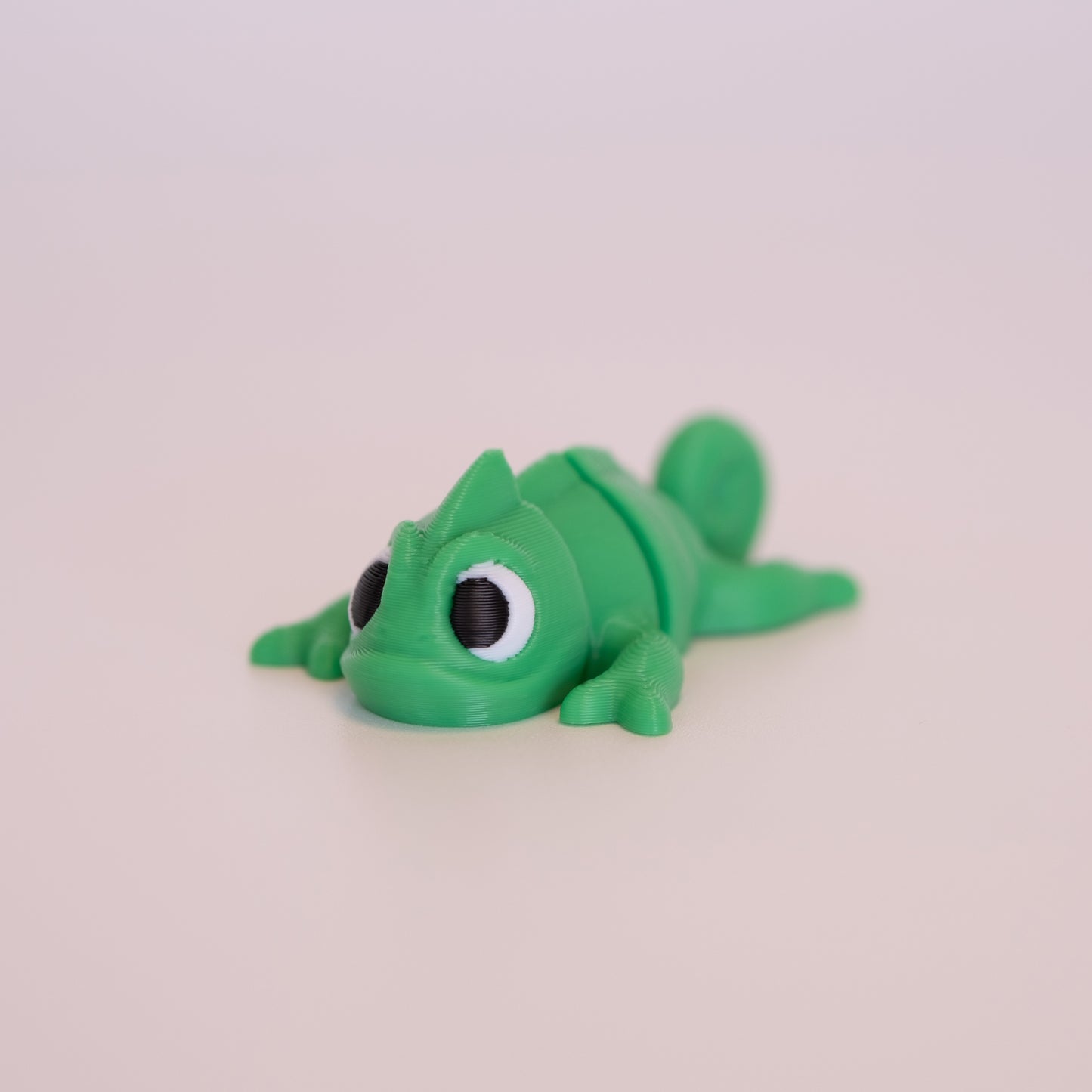 A 3D printed Chameleon figurine laying on its stomach over an off white background. The figurine is mostly green with round black eyes,white scleras, and black iris/pupils looking forward.