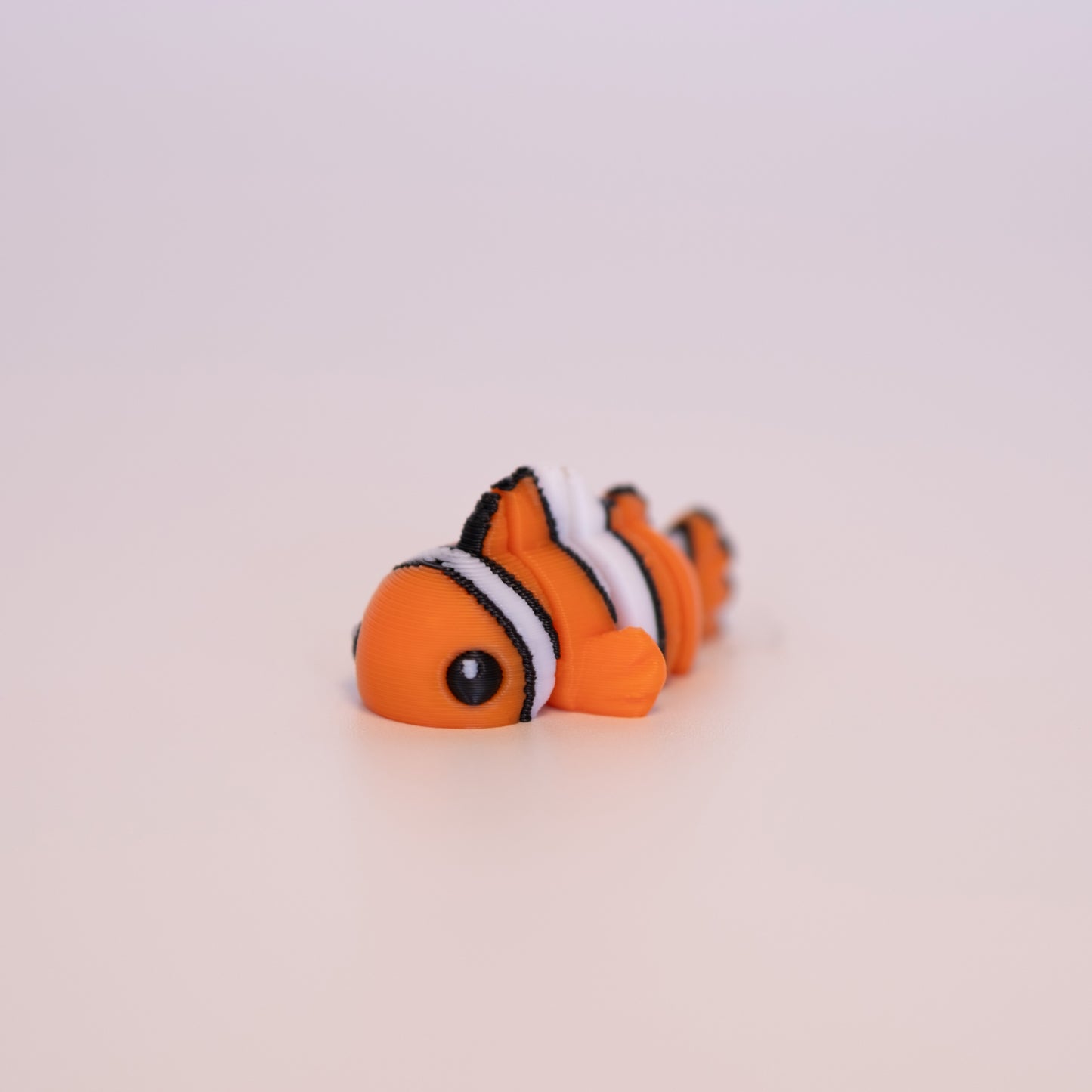 A 3D printed Clown Fish figurine laying on its stomach over an off white background. The figurine is orange with white and black striped accents. It also has round black eyes with a small white accent dot in each eye.