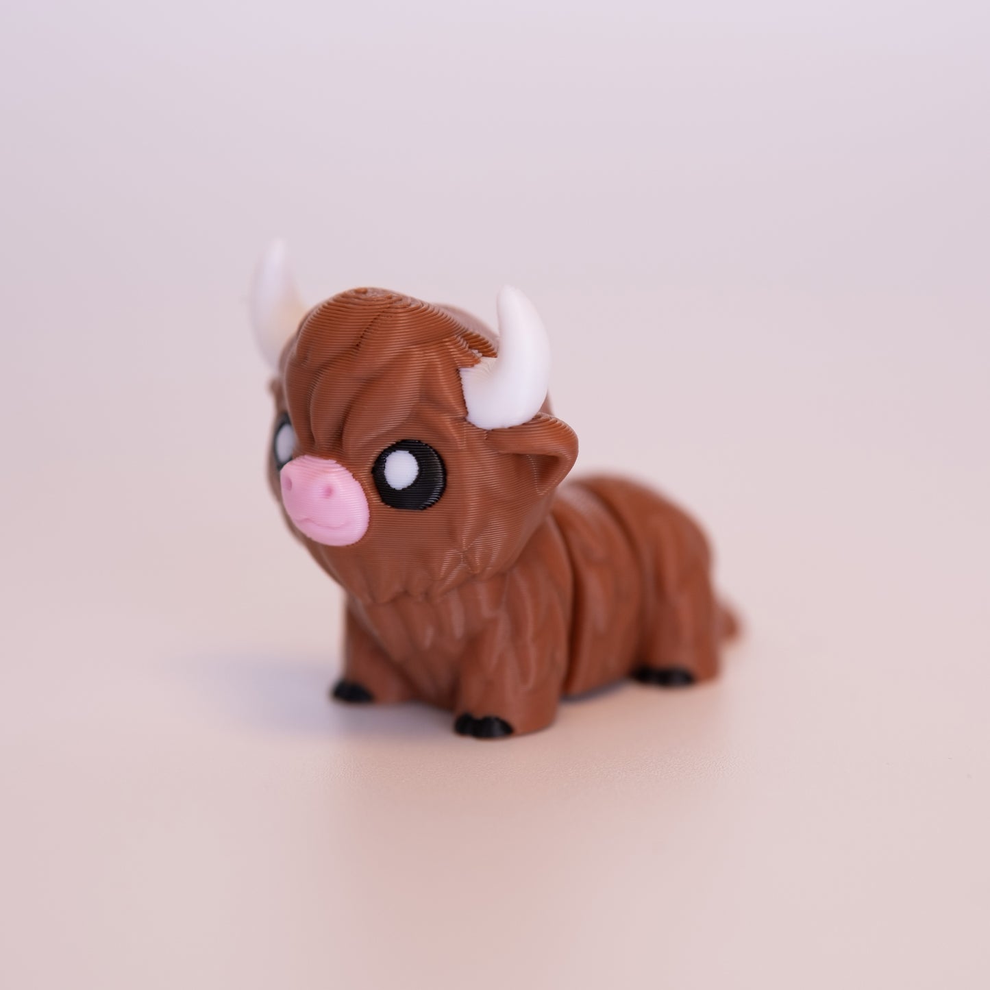 A 3D Printed Highland Cow figurine standing on its 4 legs on an off white background. The Highland Cow is mostly a warm brown, with white horns, pink nose, and black hooves. The cow also has round black eyes with 1 white accent dot in each eye.