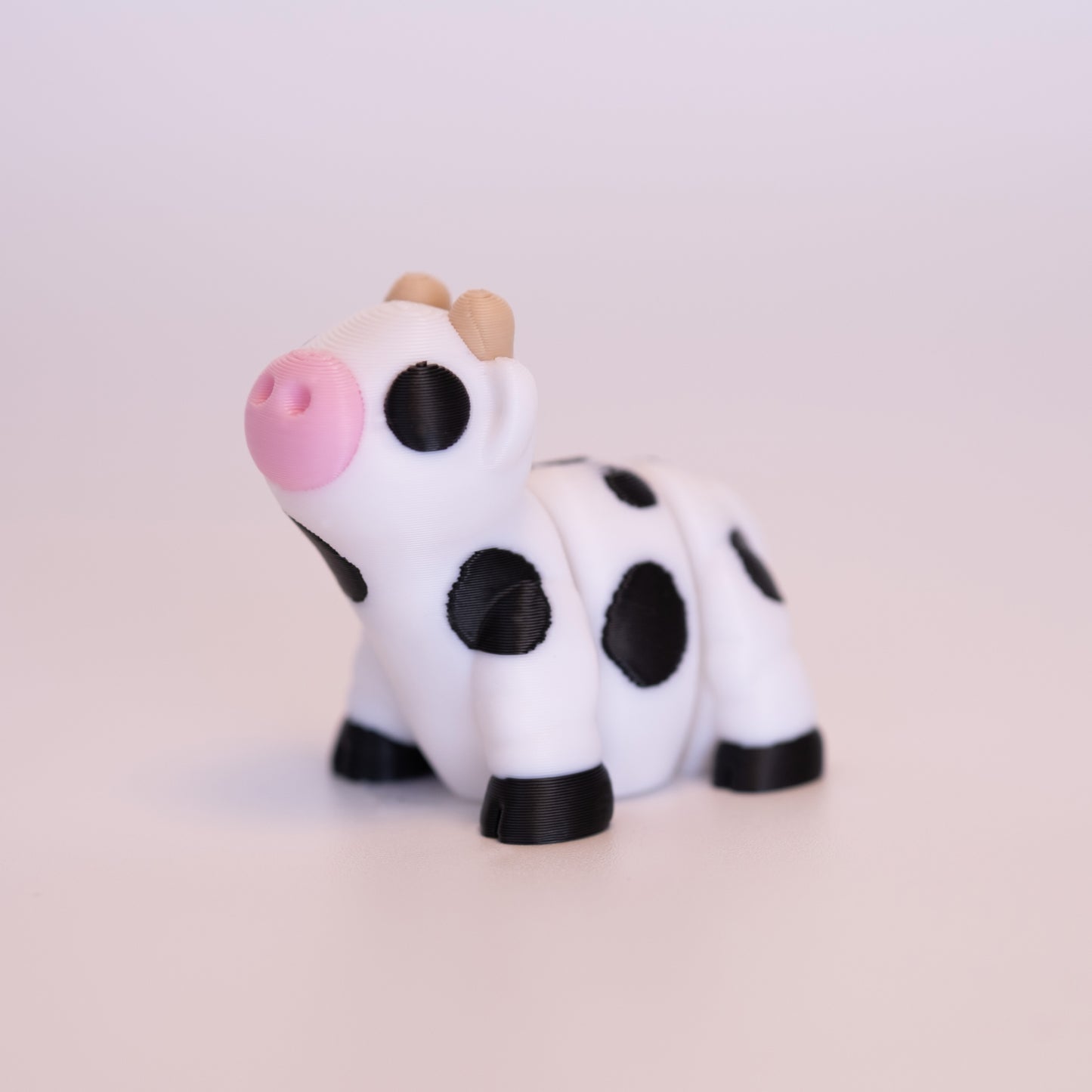 A 3D Printed Cow figurine standing on its 4 legs on an off white background. The Cow is mostly white, with round black eyes, pink nose, tan horns, black hooves, and black spotted accents.