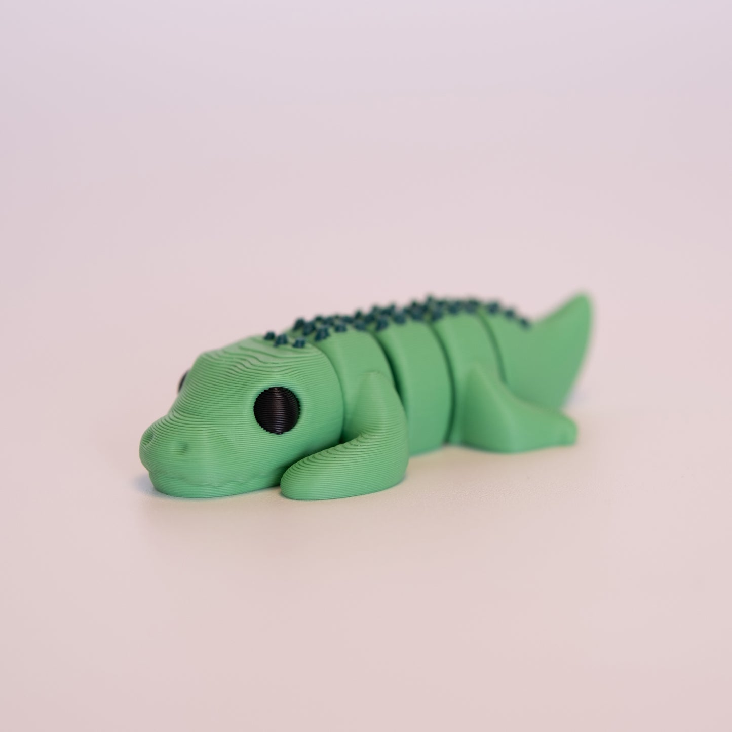 A 3D printed Crocodile figurine laying on its stomach over an off white background. The figurine is entirely green with round black eyes, and dark green accents.