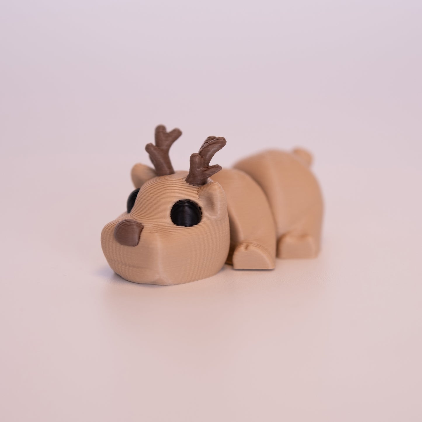 A 3D printed Deer figurine laying on its stomach over an off white background. The figurine is entirely brown with round black eyes, brown nose, and brown antlers.