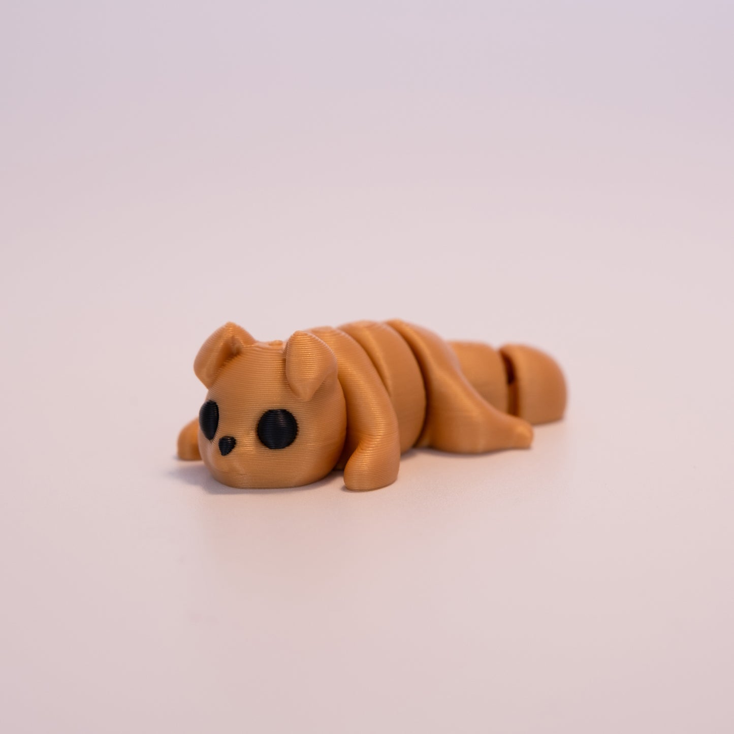 A 3D printed Gold Dog figurine laying on its stomach over an off white background. The figurine is entirely gold with round black eyes and a black button nose.