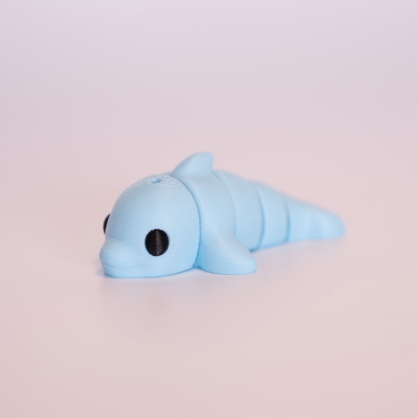 A 3D printed Dolphin figurine laying on its stomach over an off white background. The figurine is entirely light blue with round black eyes.