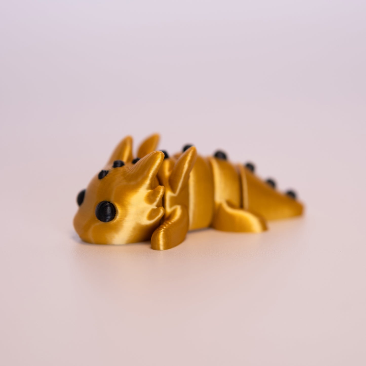 A 3D printed Gold Dragon figurine laying on its stomach over an off white background. The figurine is entirely gold with round black eyes and black accents.