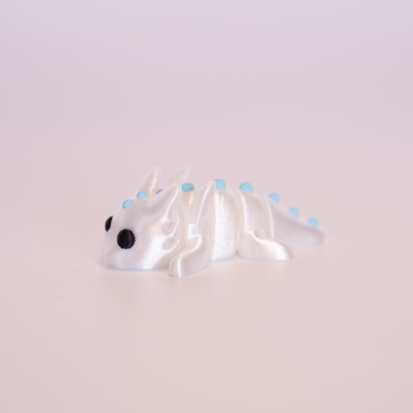 A 3D printed White Dragon figurine laying on its stomach over an off white background. The figurine is entirely shiny white with round black eyes and light blue accents.
