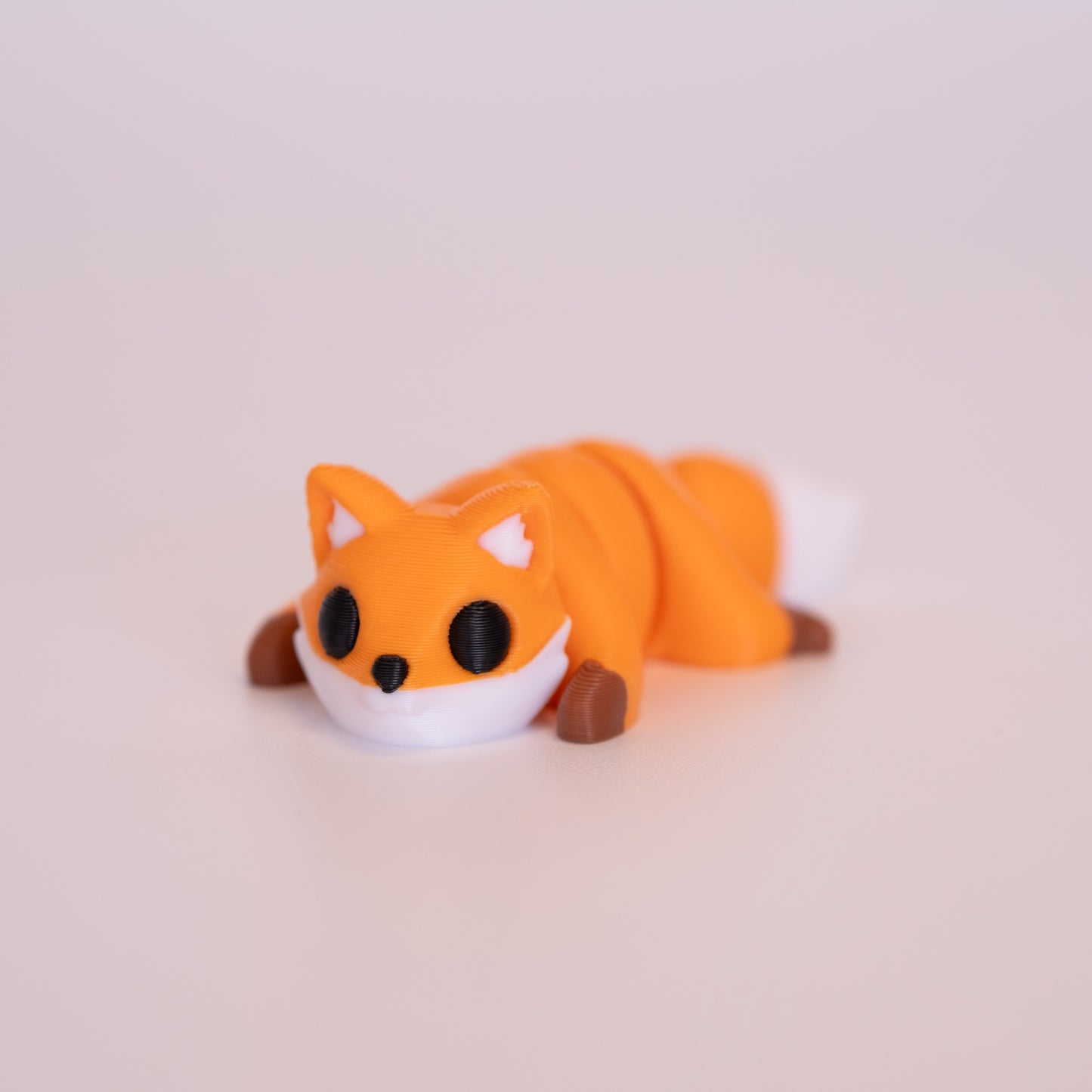 A 3D printed Red Fox figurine laying on its stomach over an off white background. The figurine is mostly orange with round black eyes, white accents, and brown paws.