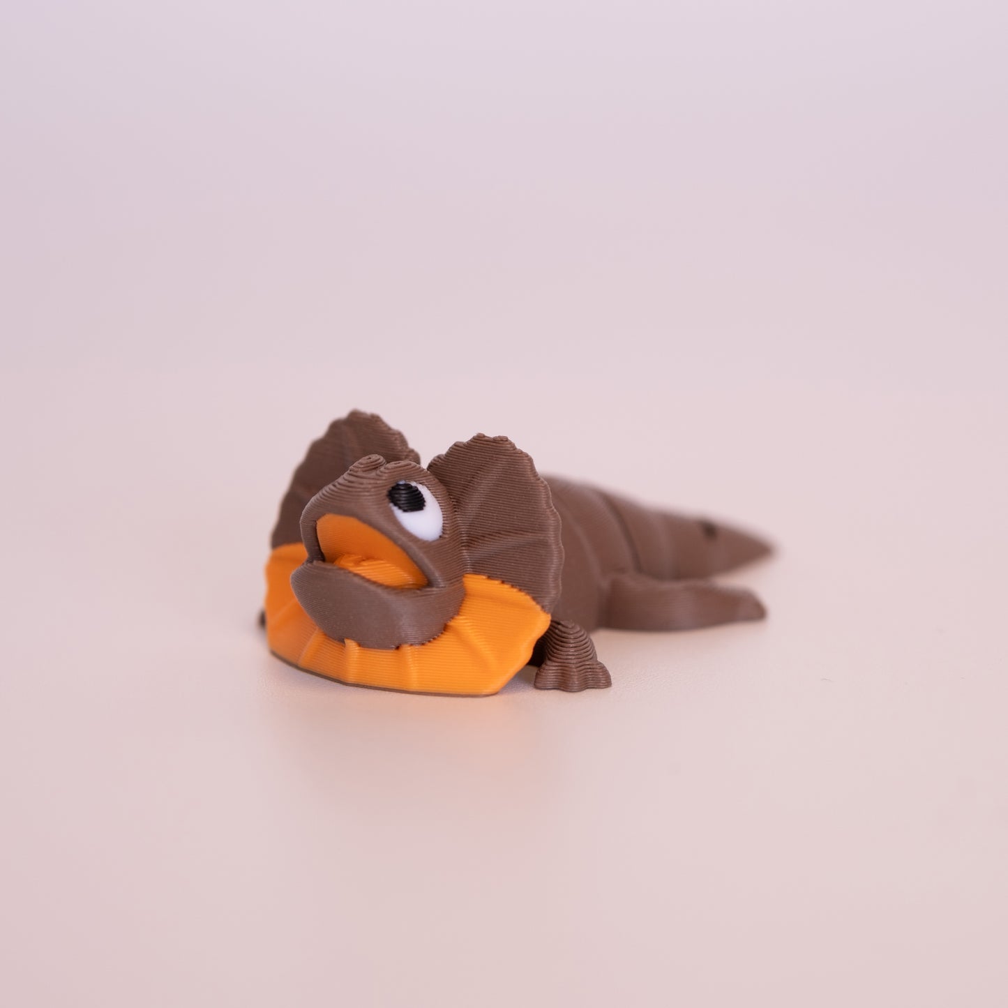 A 3D printed Frilled Neck Lizard figurine laying on its stomach over an off white background. The figurine is mostly brown, orange accents, white scleras, and black iris/pupils looking up.