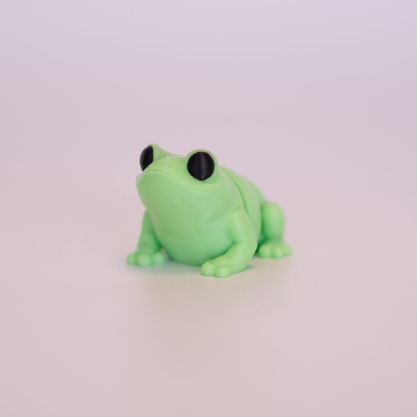 A 3D Printed Frog figurine standing on its 4 legs on an off white background. The Frog is mostly light green with round black eyes.