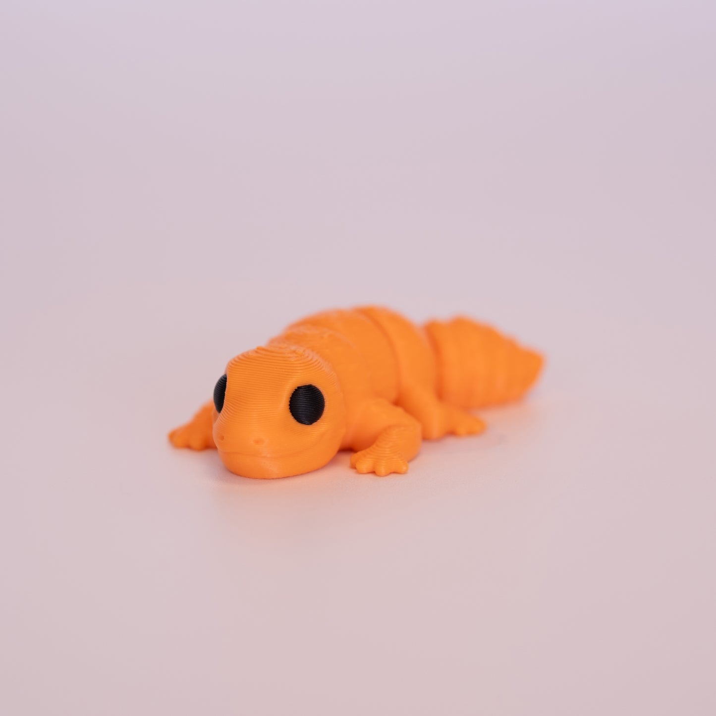 A 3D printed Gecko figurine laying on its stomach over an off white background. The figurine is entirely orange with round black eyes.