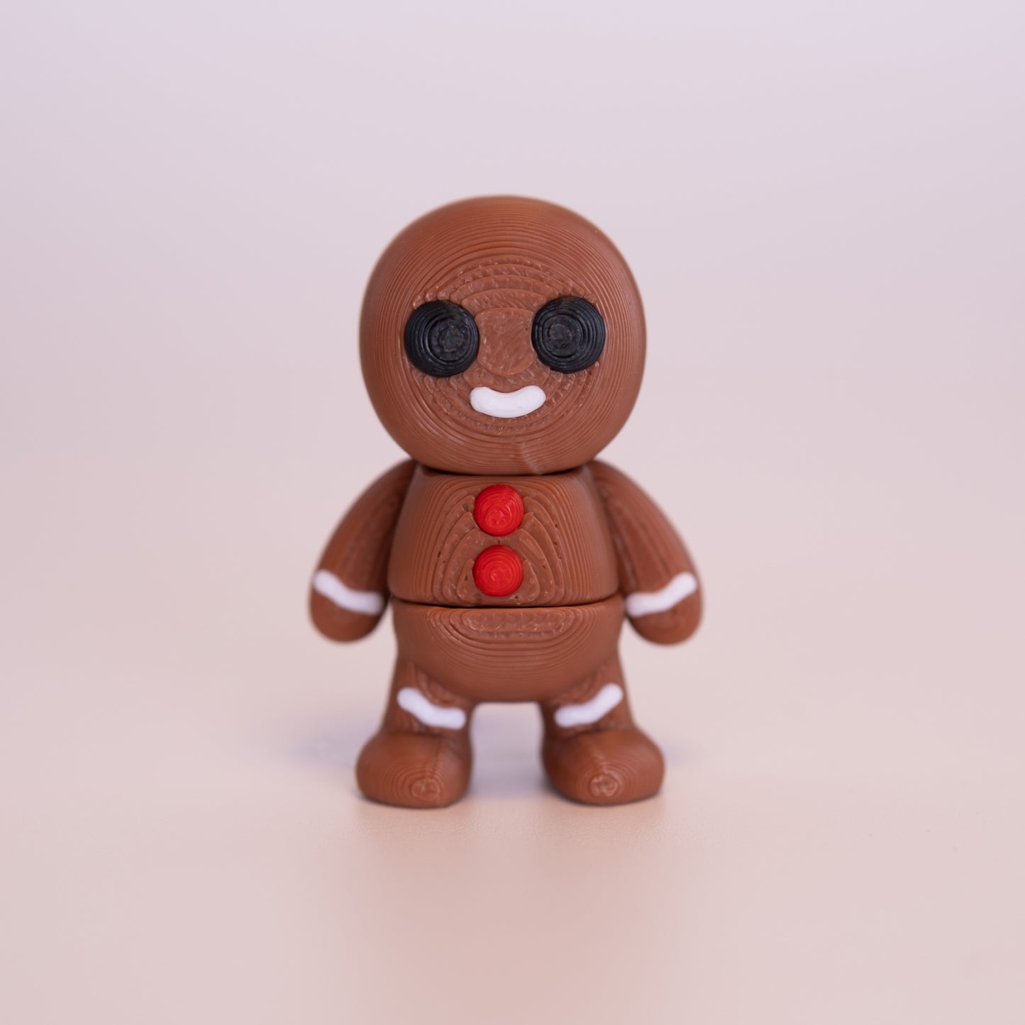 A 3D printed standing Gingerbread Man figurine on an off white background. The Gingerbread Man is a mostly warm brown, with round black eyes, red buttons, white smile, and additional white accents.