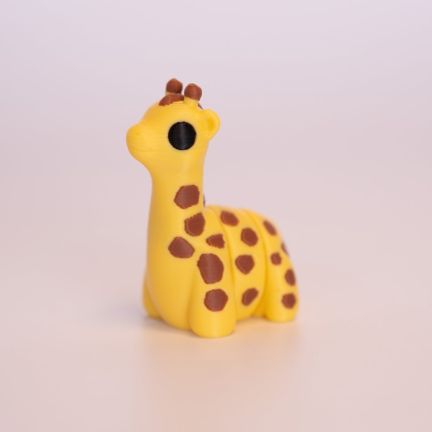 A 3D Printed Giraffe figurine standing on its 4 legs on an off white background. The Giraffe is mostly yellow with round black eyes, brown ossicones, and brown spotted accents all over the body.
