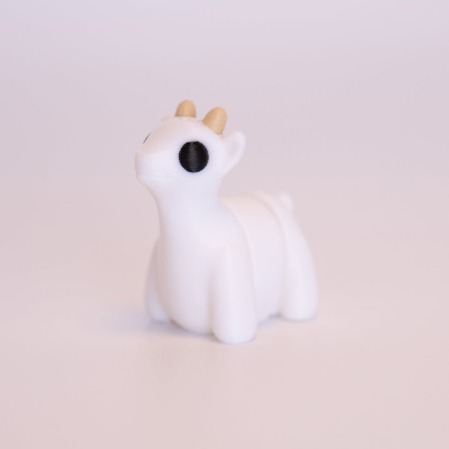 A 3D Printed Goat figurine standing on its 4 legs on an off white background. The Goat is mostly white with round black eyes, and beige horns.