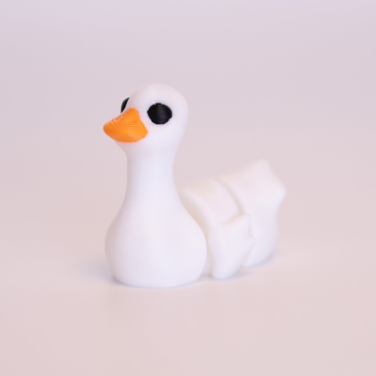 A 3D printed Goose sitting on an off white background. The Goose is mostly white with black eyes, and an orange beak.