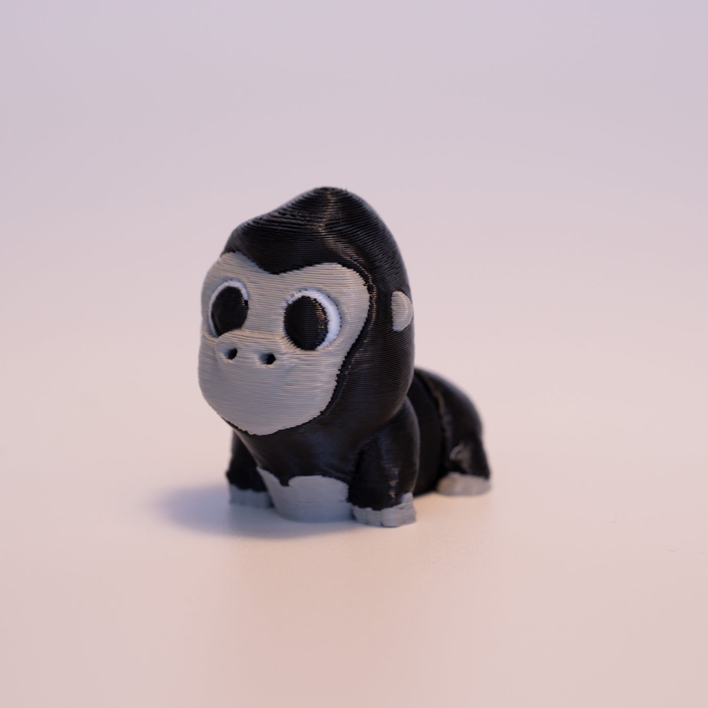A 3D Printed Gorilla figurine standing on its 4 legs on an off white background. The Gorilla is mostly black and has grey accents on the face, body, ears, feet, and hands. Its round black eyes have white scaleras surrounding them.