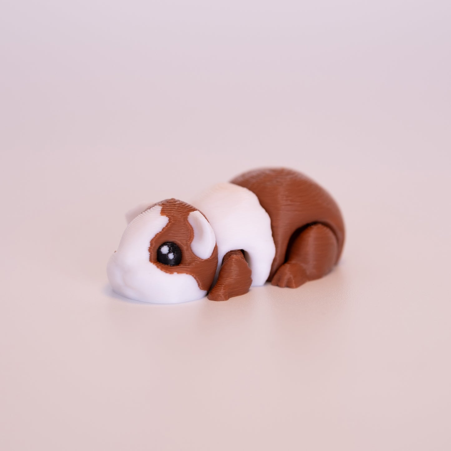 A 3D printed Guinea Pig figurine laying on its stomach over an off white background. The figurine is mostly a warm brown with white accents. The eyes are round and black with 2 white accent dots in each eye.