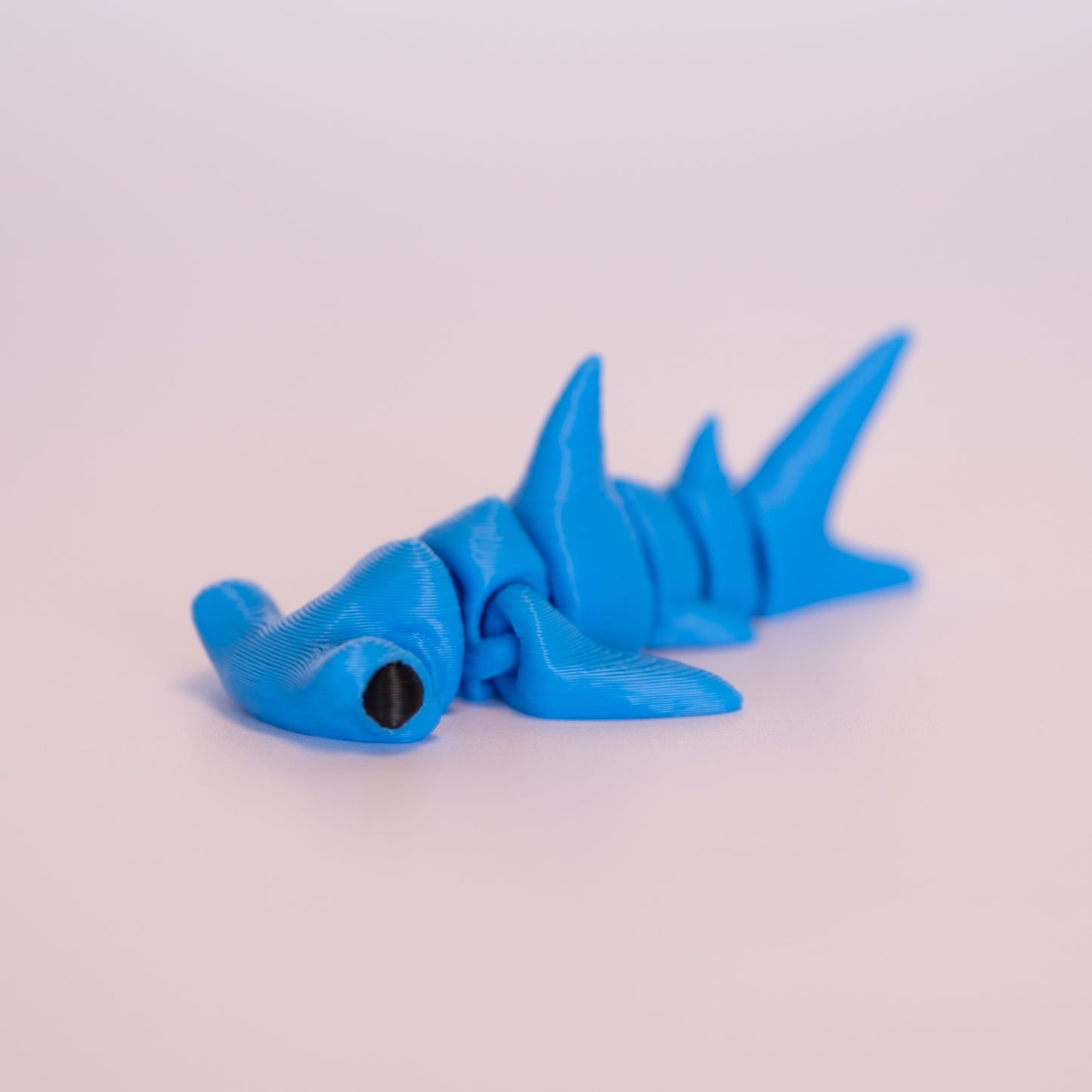 A 3D printed Hammerhead Shark figurine laying on its stomach over an off white background. The figurine is entirely blue with round black eyes.