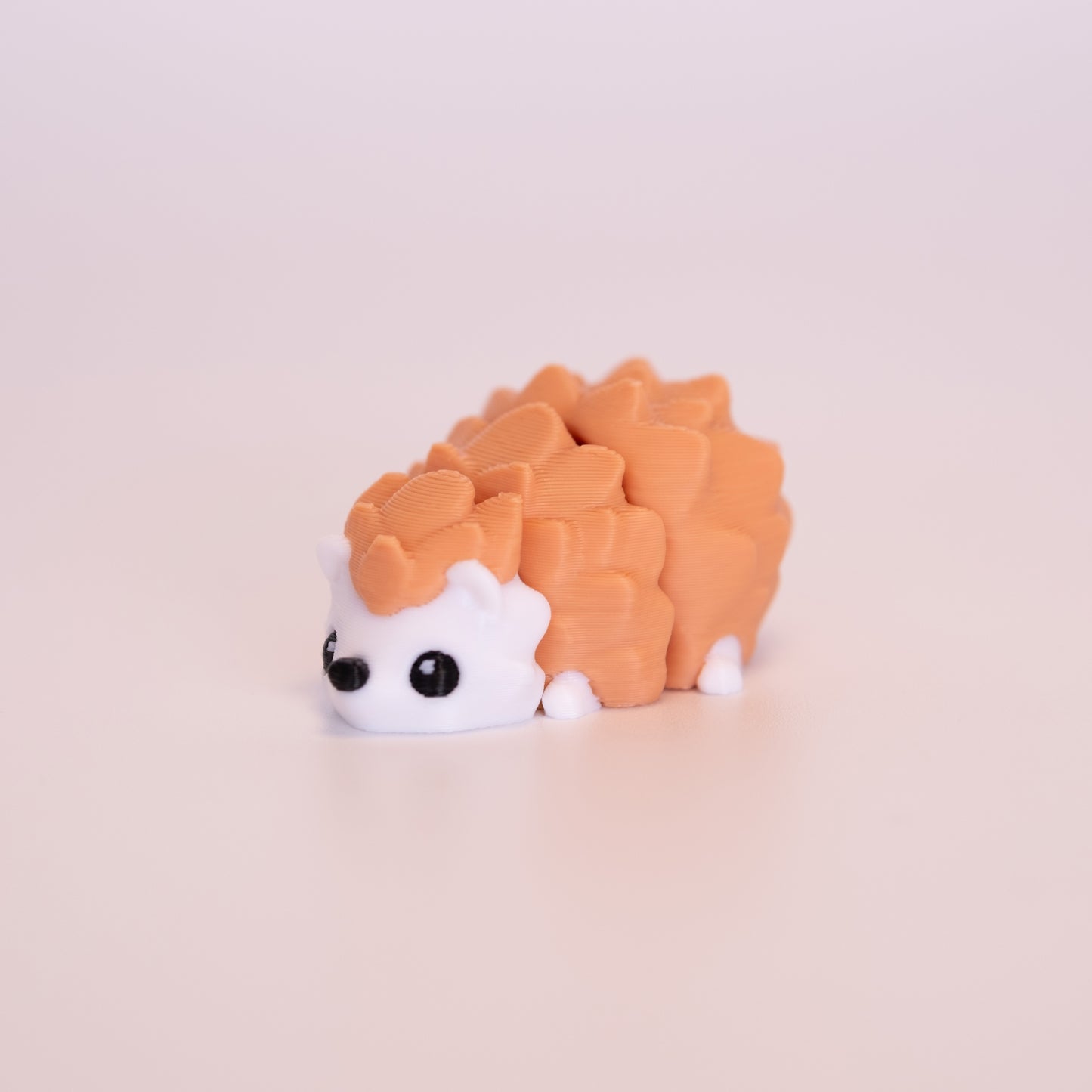 A 3D printed Hedgehog figurine laying on its stomach over an off white background. The figurine has muted orange pins, a white body, black nose, and black eyes that have one white accent dot in each eye.