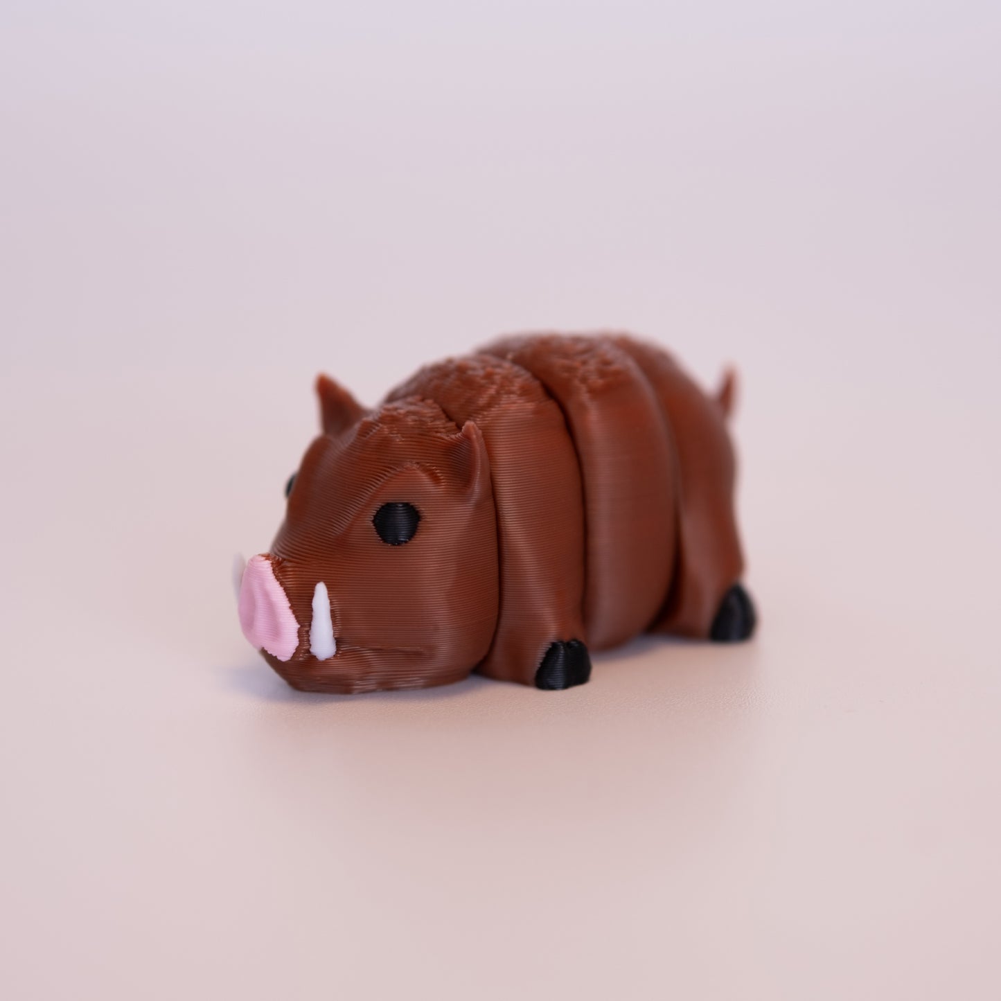 A 3D printed Hog figurine laying on its stomach over an off white background. The figurine is mostly a warm brown with round black eyes, pink nose, white tusks, and black hooves.