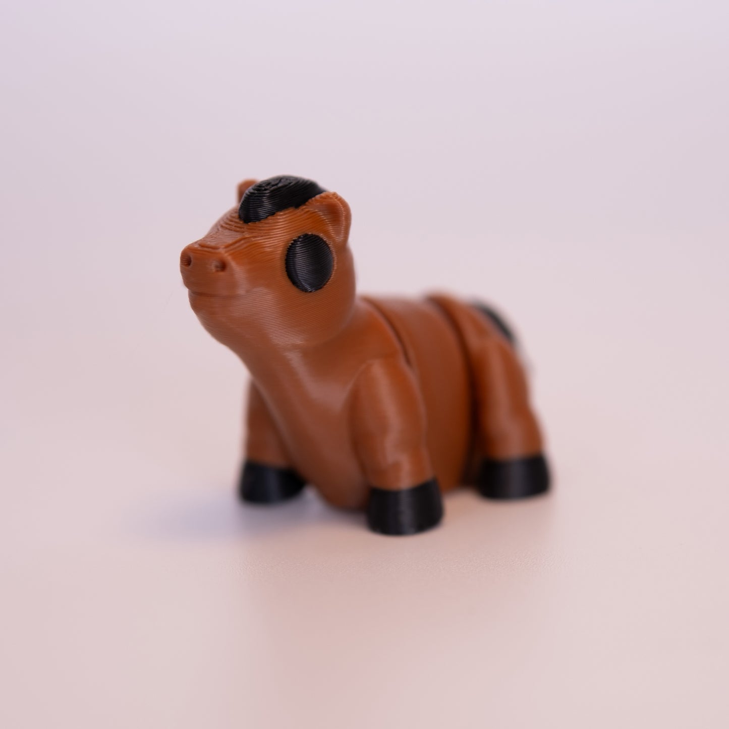 A 3D Printed Horse figurine standing on its 4 legs on an off white background. The Horse is mostly a warm brown with round black eyes, black hooves, black tail, and black mane.