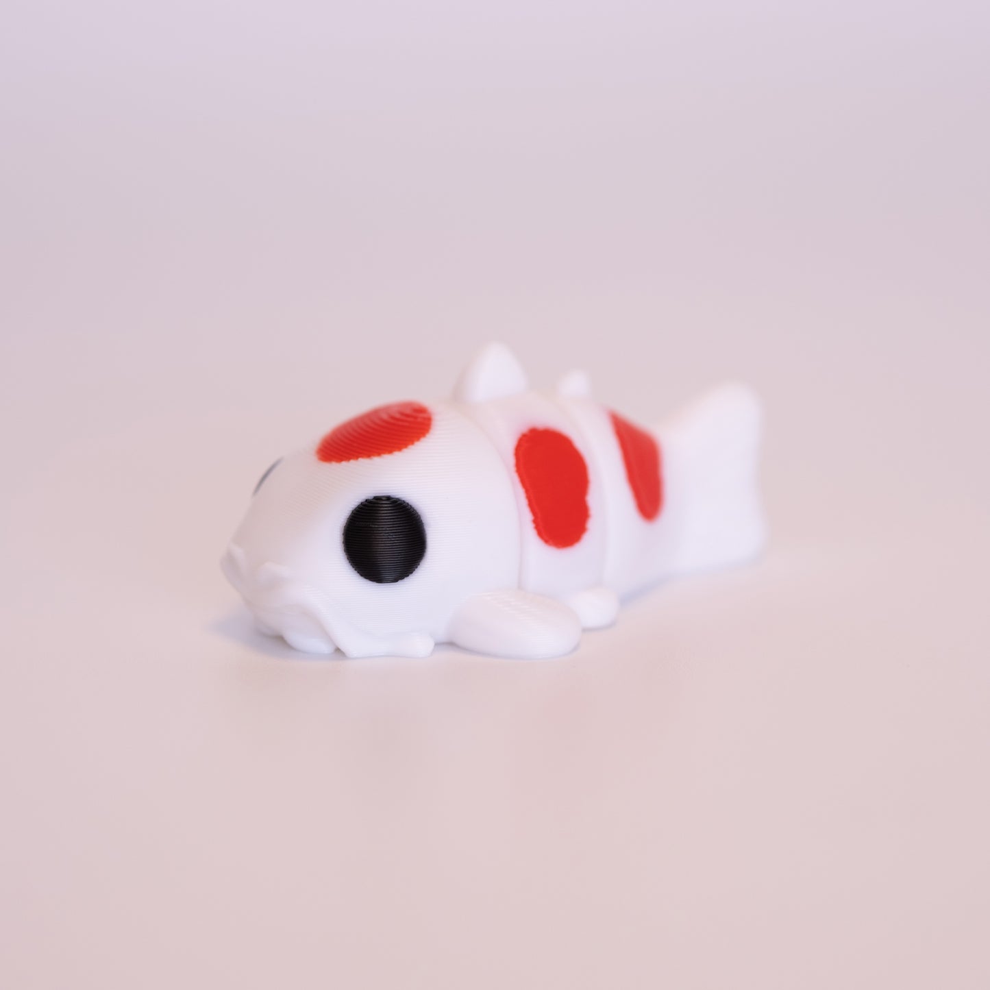A 3D printed Koi Fish figurine laying on its stomach over an off white background. The figurine is mostly white with round black eyes and red accents.