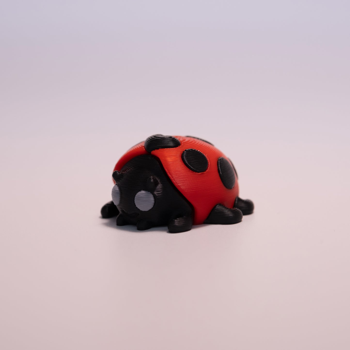 A 3D printed LadyBug figurine laying on its stomach over an off white background. The figurine is mostly black with round grey eyes, red wings, and black accent dots.