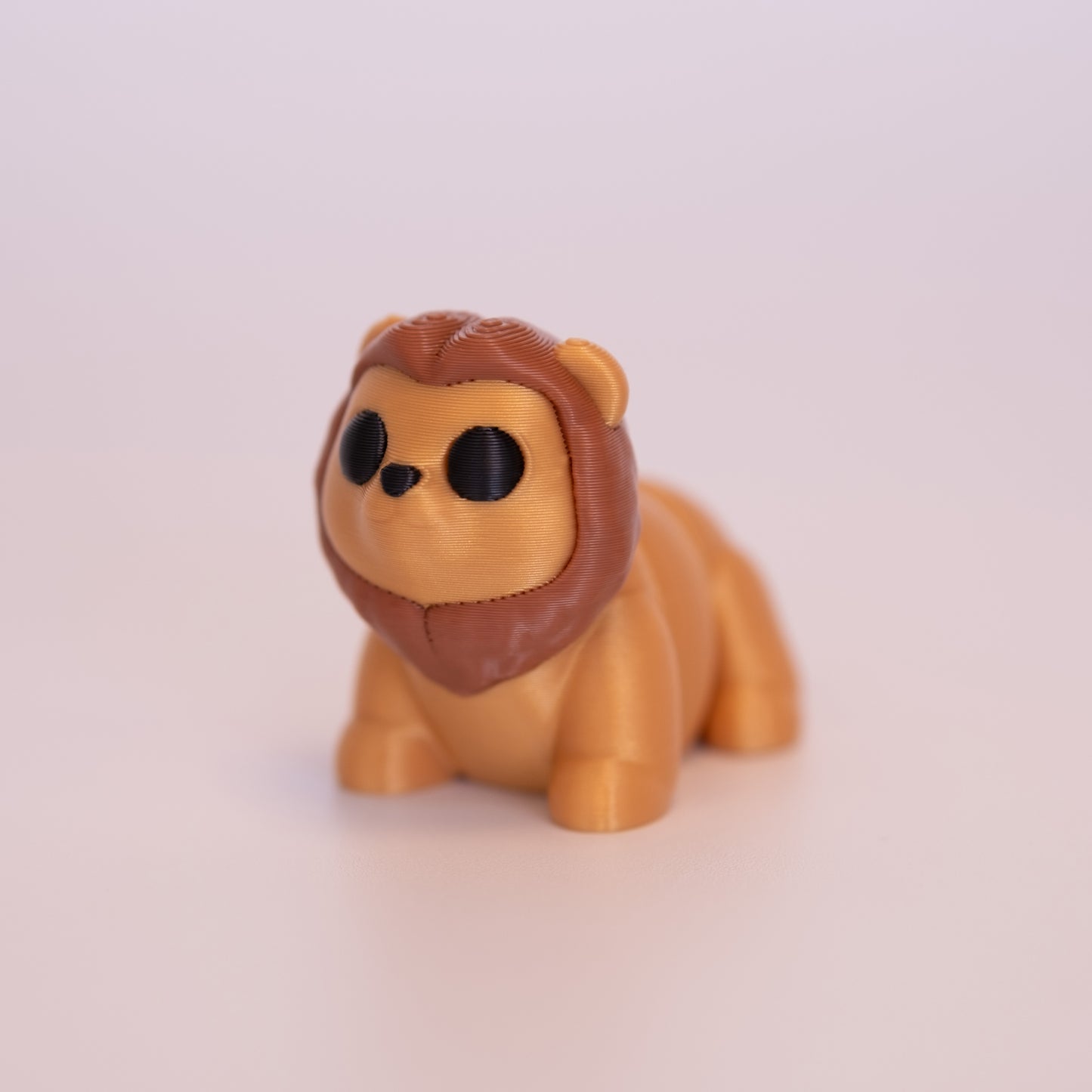 A 3D Printed Lion figurine standing on its 4 legs on an off white background. The Lion is mostly gold, with a brown mane, round black eyes, and a black nose.