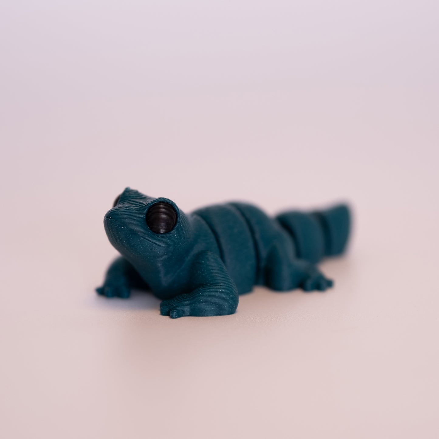 A 3D printed Lizard figurine laying on its stomach over an off white background. The figurine is entirely green with round black eyes.