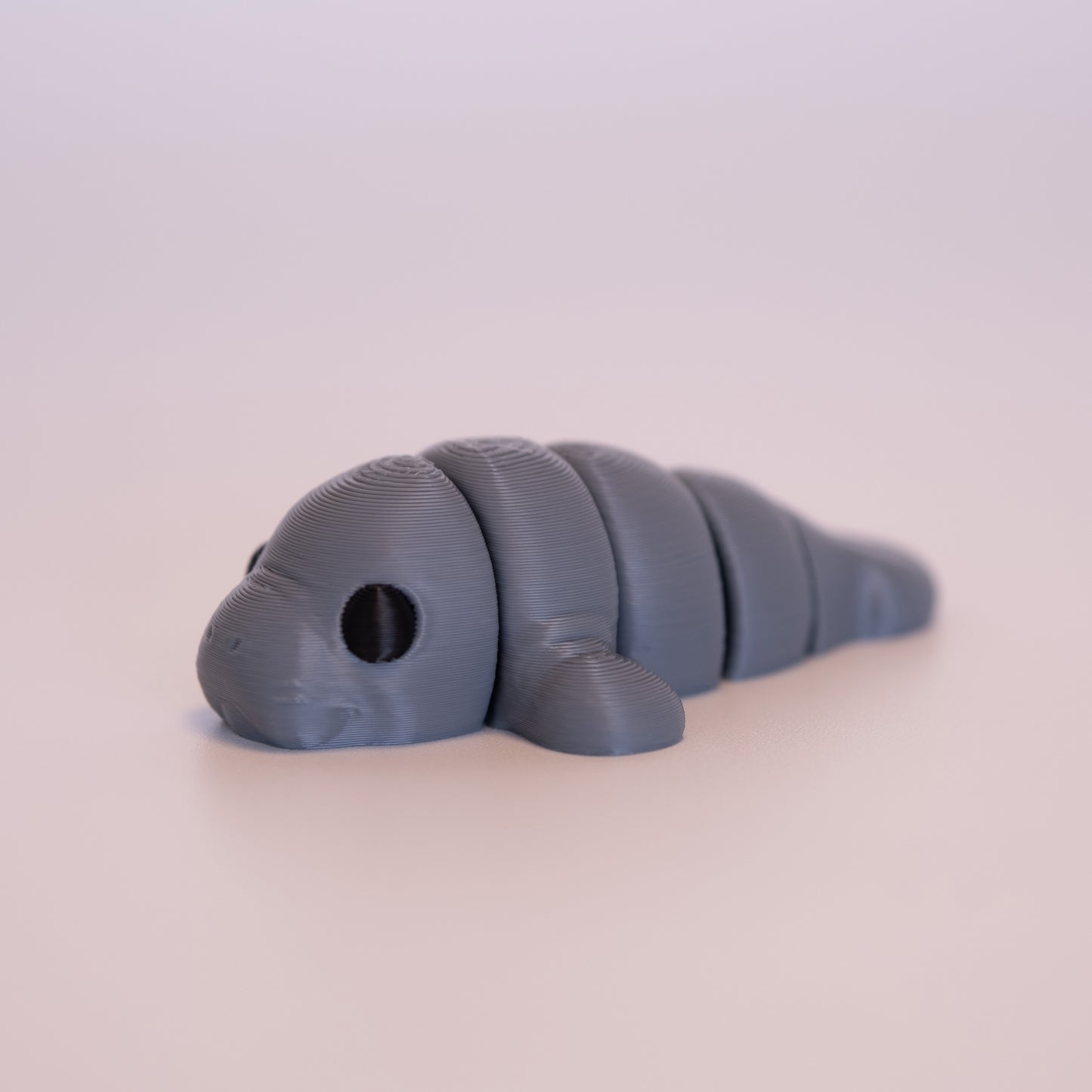 A 3D printed Manatee figurine laying on its stomach over an off white background. The figurine is entirely grey with round black eyes.