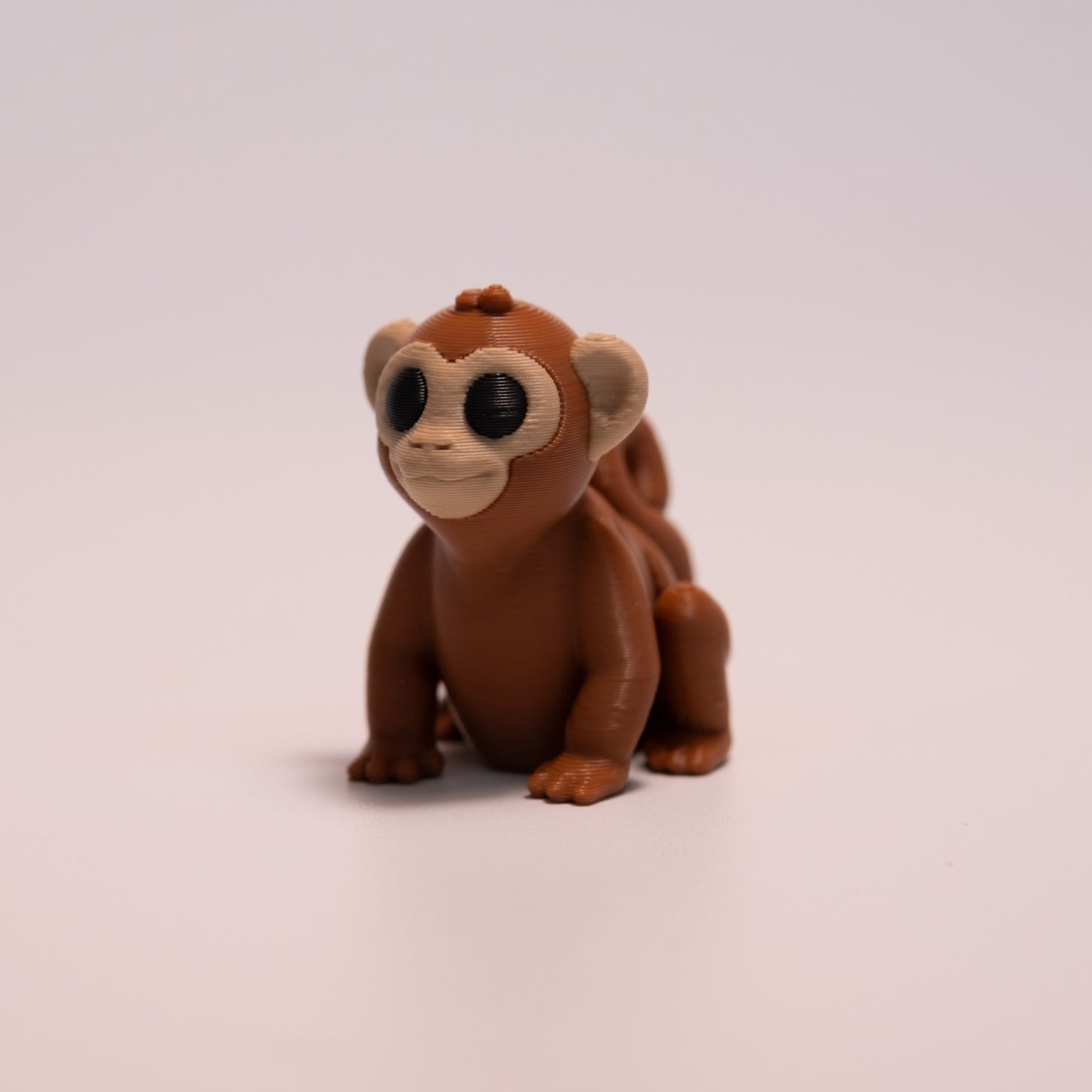 A 3D Printed Monkey figurine standing on its 4 legs on an off white background. The Monkey is mostly a warm brown with round black eyes, and tan accents on the face and ears.