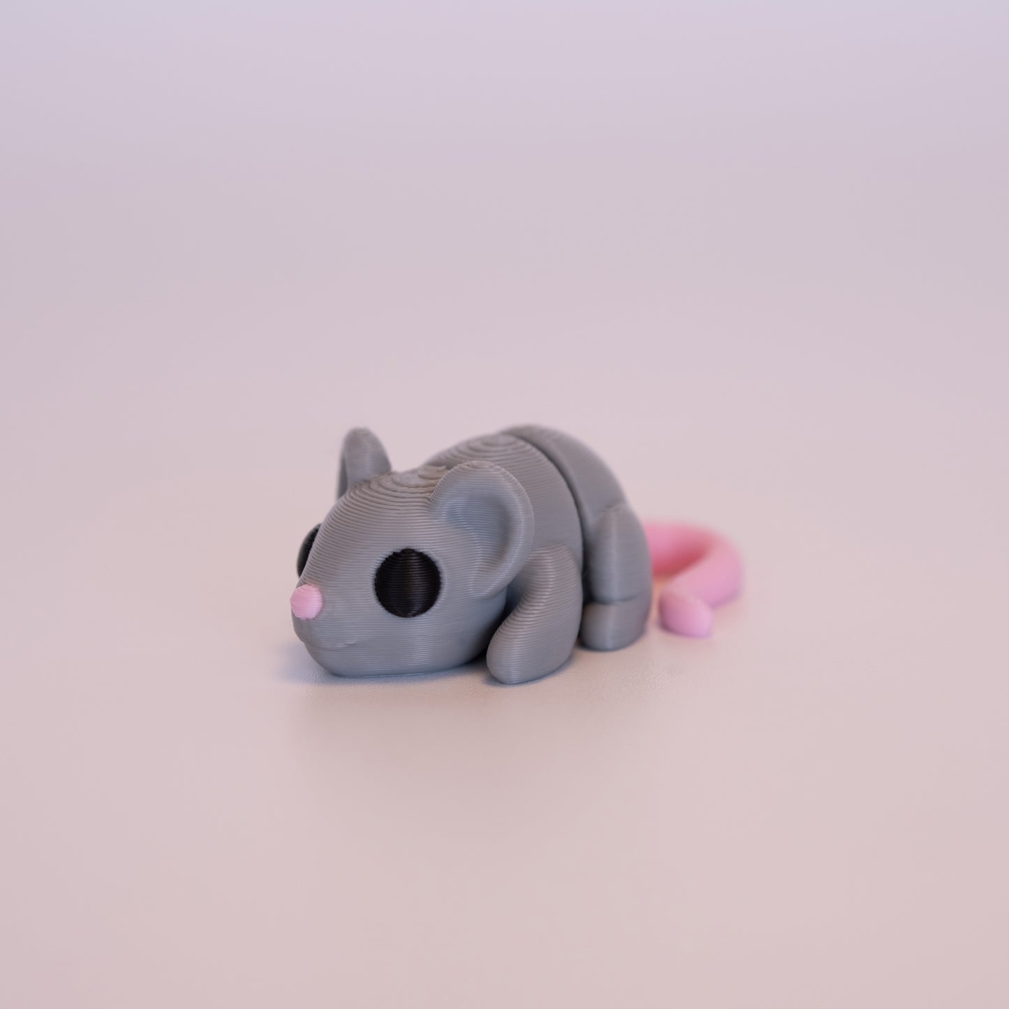 A 3D printed Mouse figurine laying on its stomach over an off white background. The figurine is mostly light grey with round black eyes, pink tail, and pink nose.