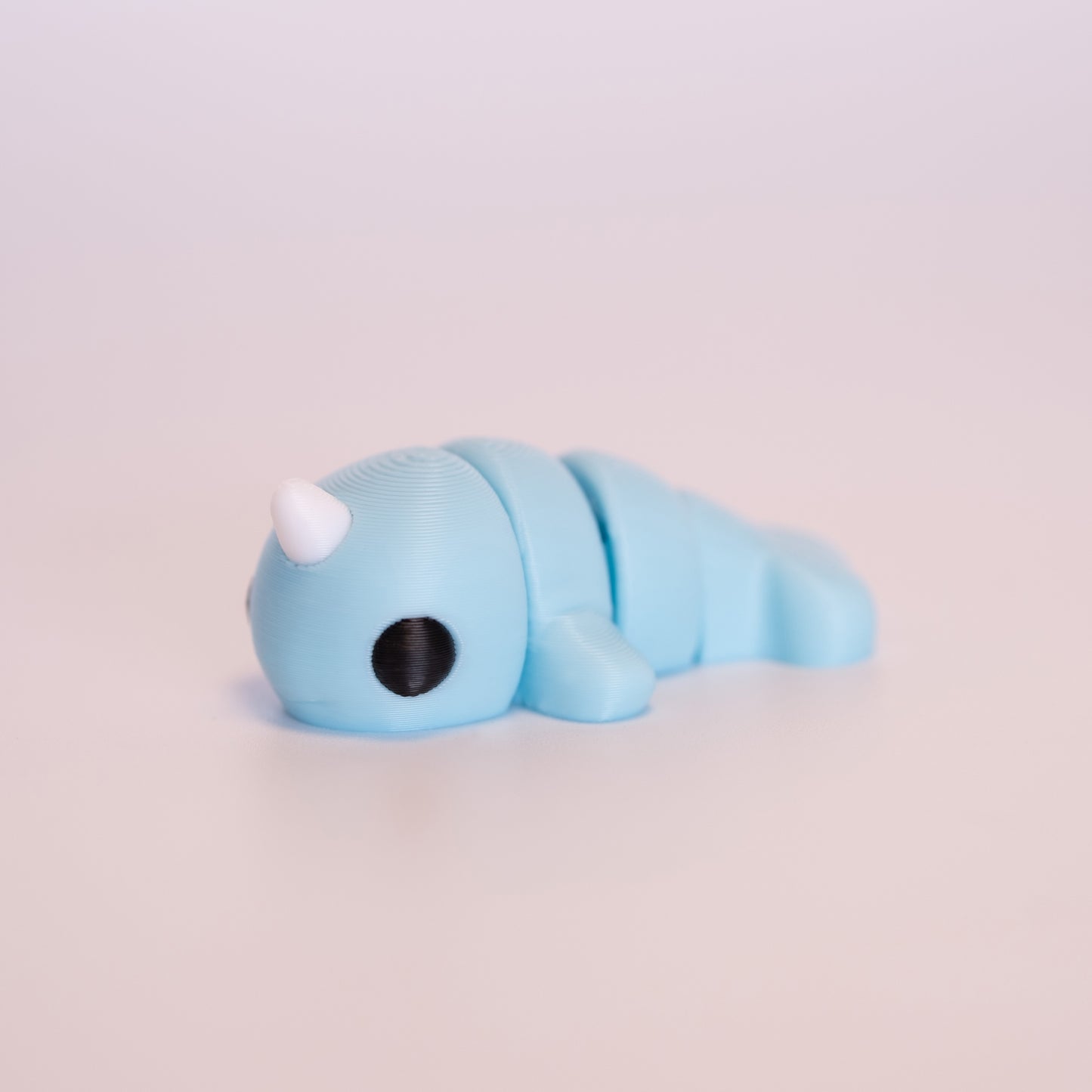 A 3D printed Narwhal figurine laying on its stomach over an off white background. The figurine is mostly light blue with round black eyes and a white horn.