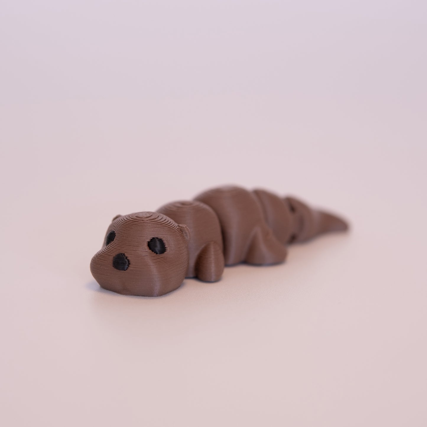 A 3D printed Otter figurine laying on its stomach over an off white background. The figurine is entirely brown with round black eyes and a black nose.