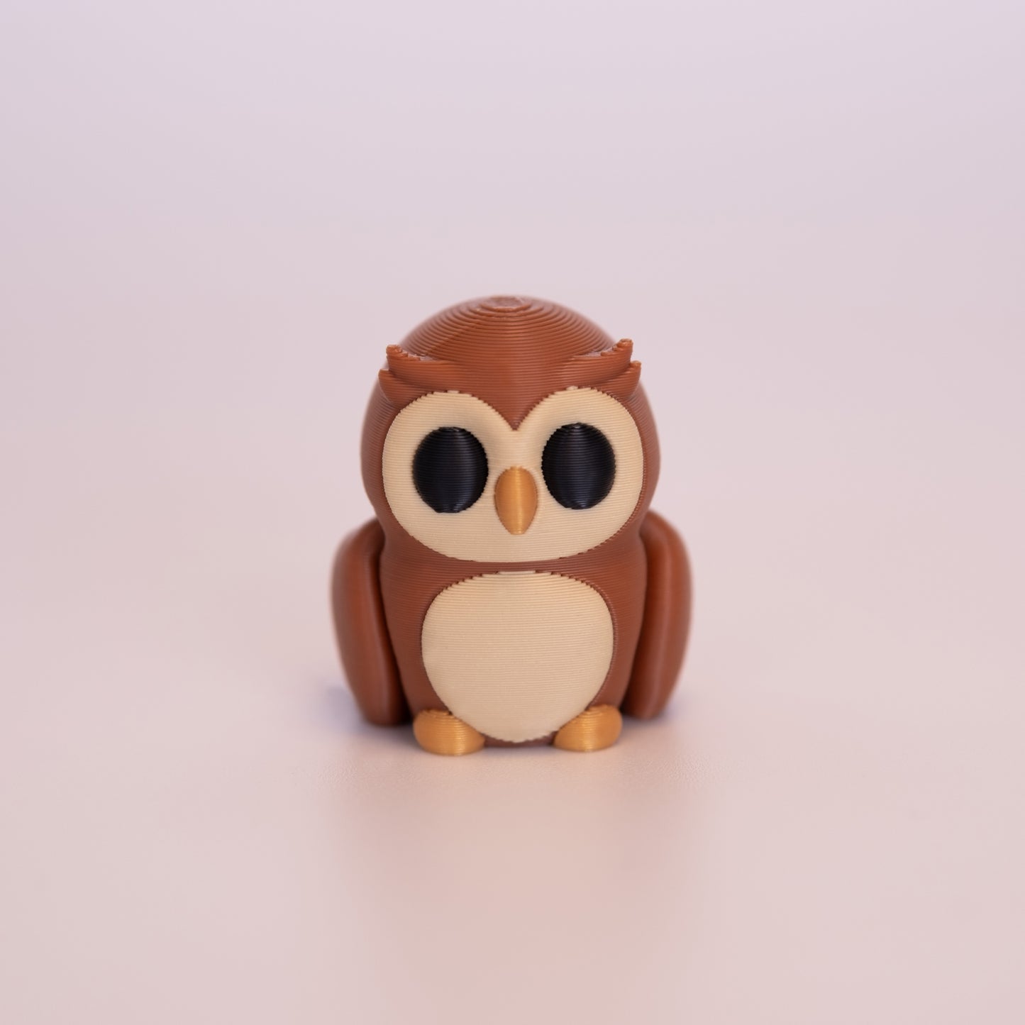 A 3D printed Owl sitting upright on an off white background. The Owl is mostly a warm brown, with round black eyes, beige accents on the face and stomach, and a gold beak and feet.