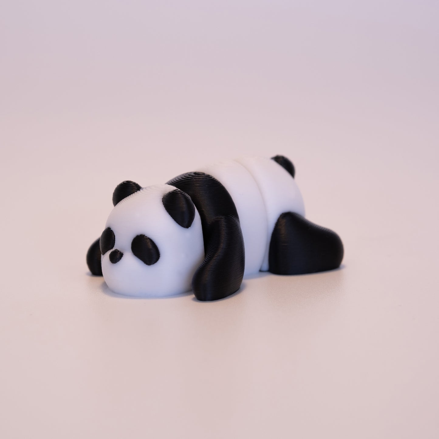 A 3D printed Panda figurine laying on its stomach over an off white background. The figurine is mostly white with black eyes, and black accents.
