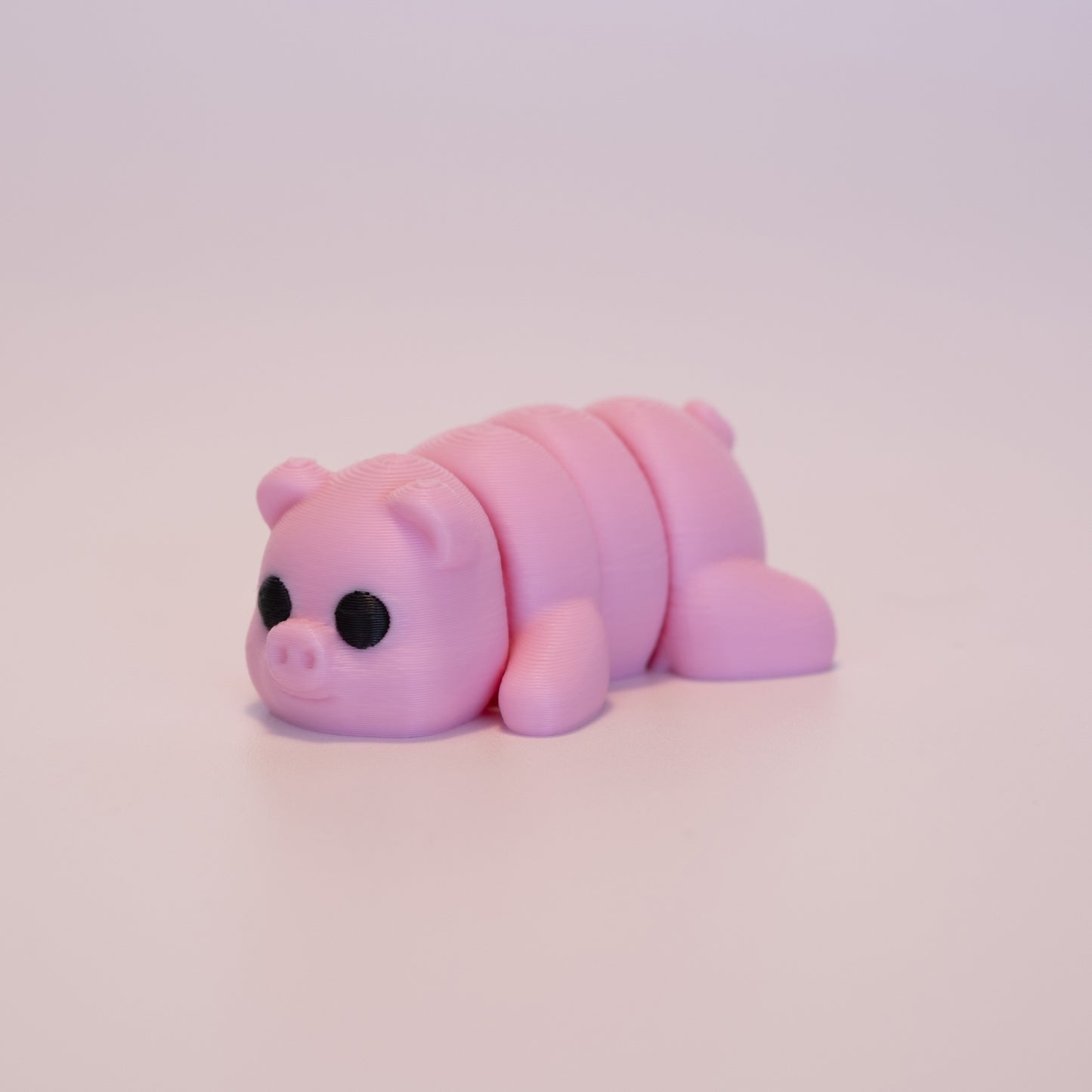 A 3D printed Pig figurine laying on its stomach over an off white background. The figurine is entirely pink with round black eyes.