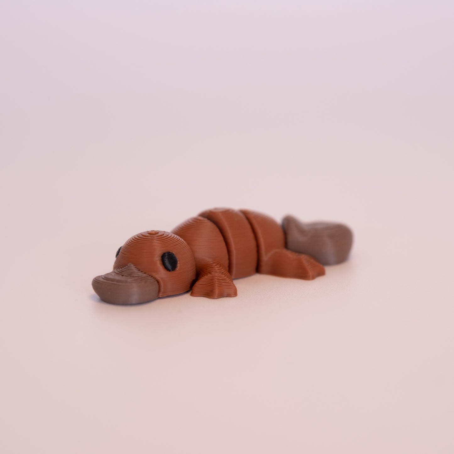 A 3D printed Platypus figurine laying on its stomach over an off white background. The figurine is mostly a warm brown and has round black eyes, with a cool brown beak and tail.