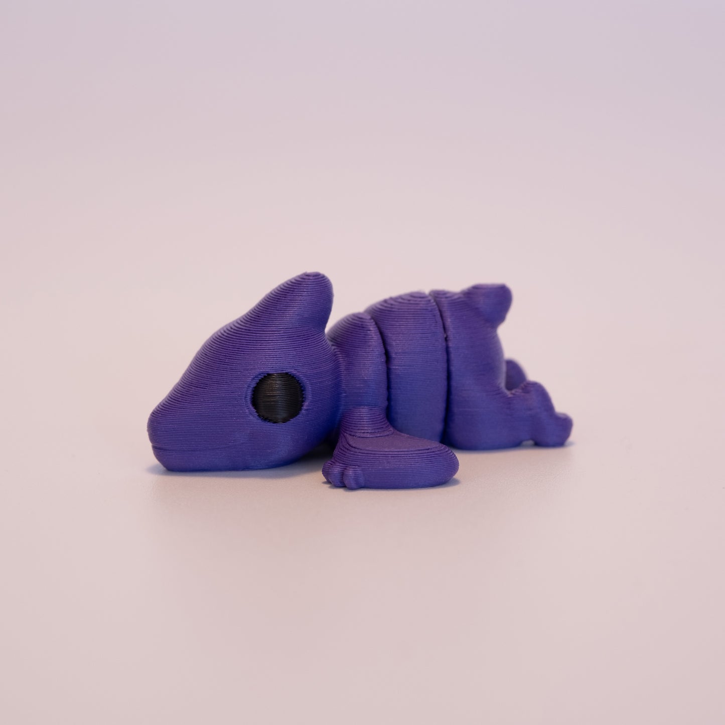A 3D printed Pterodactyl figurine laying on its stomach over an off white background. The figurine is entirely dark purple with round black eyes.